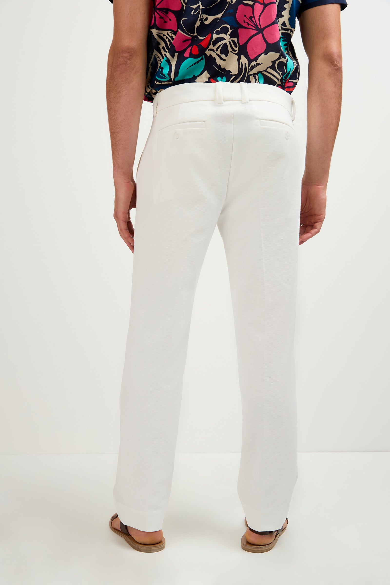 A person stands facing away, wearing Clyde Slim Trouser - Stretch Piqué in white, paired with a colorful floral shirt and sandals against a plain white background.