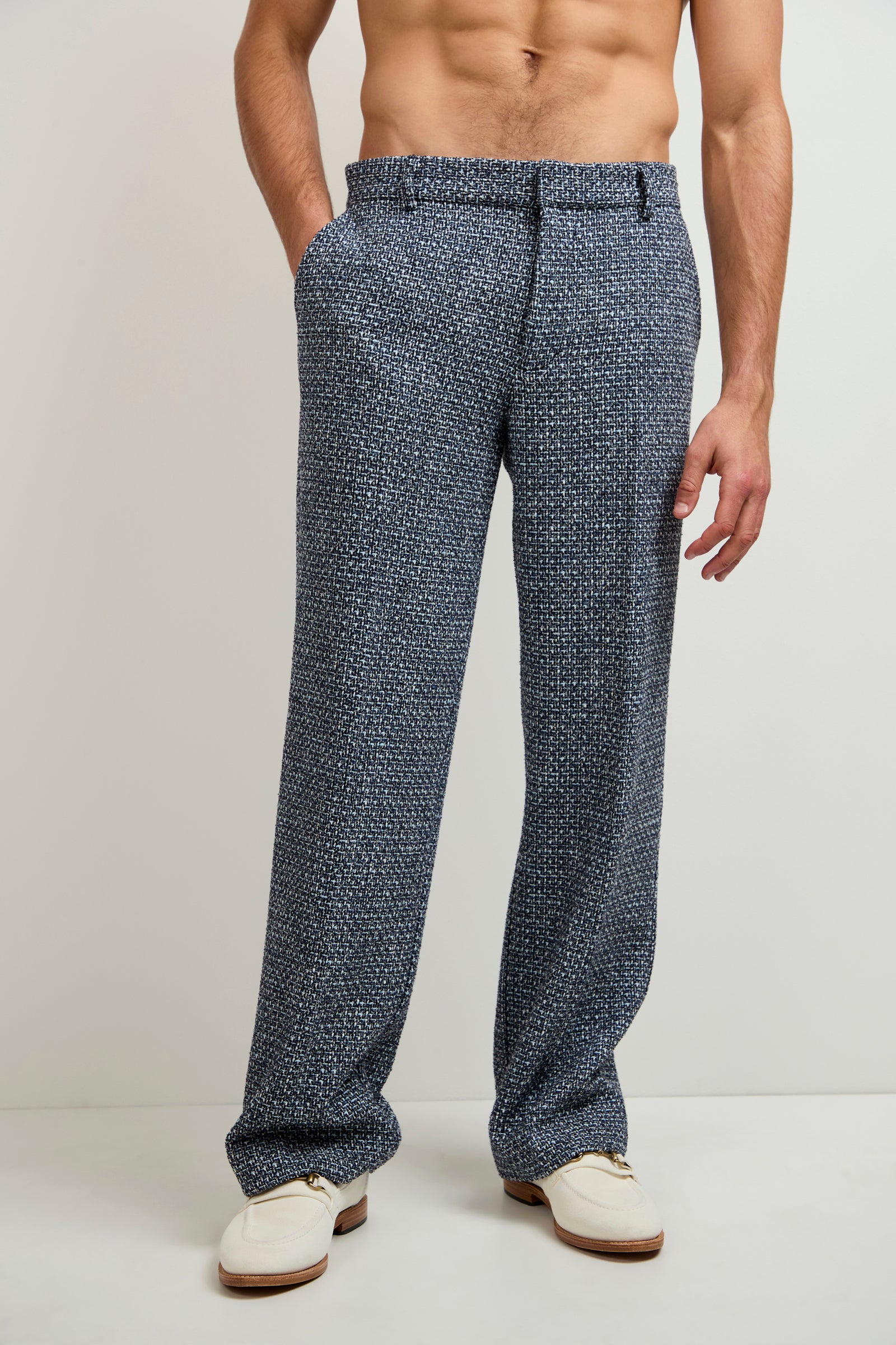 A shirtless man stands against a plain, light-colored background with his hands in his pockets, wearing Buckingham Tweed Trouser in Selva Suiting and white shoes.