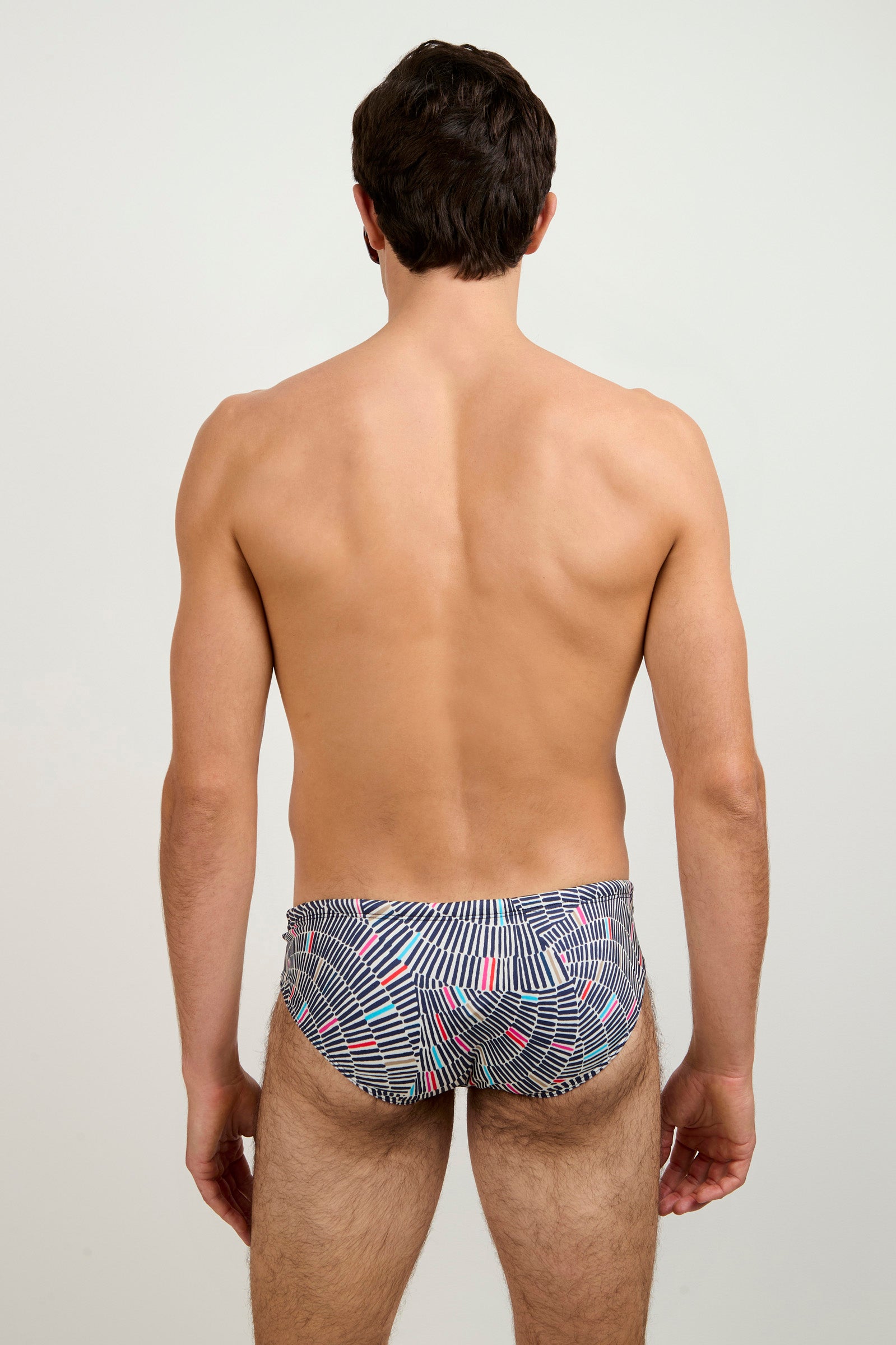 A man faces away from the camera, showing his back and arms against a plain light background, wearing the Sorrento Swim Trunk - Java, which features a geometric black, white, and red pattern.