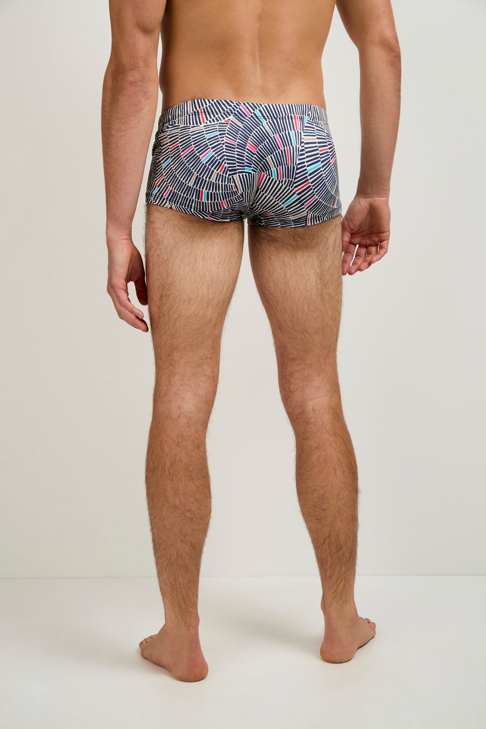 A man stands barefoot with his back to the camera, wearing the Sorrento Swim Trunk - Java, featuring black, white, and red geometric lines, set against a plain light background.