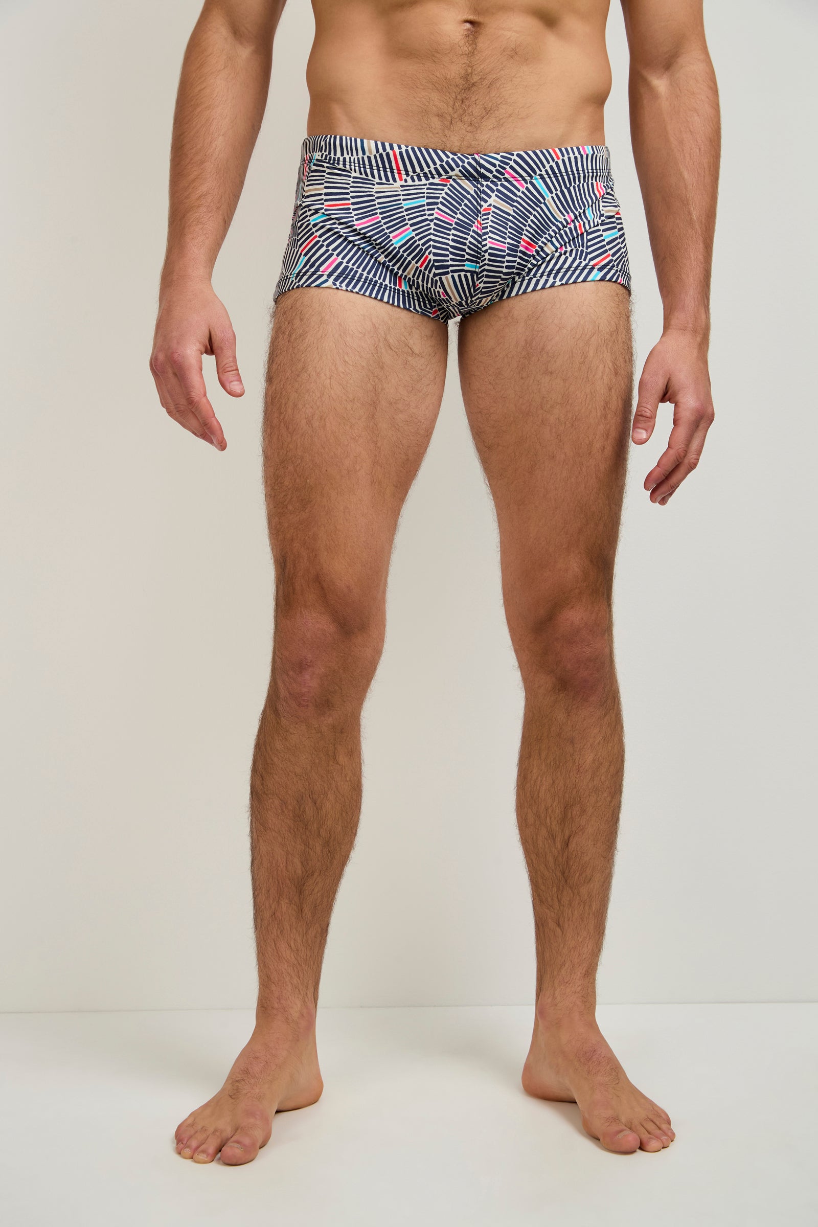 A man stands barefoot against a plain background, wearing the Sorrento Swim Trunk - Java, featuring horizontal stripes and small red accents. His upper body is mostly out of frame.