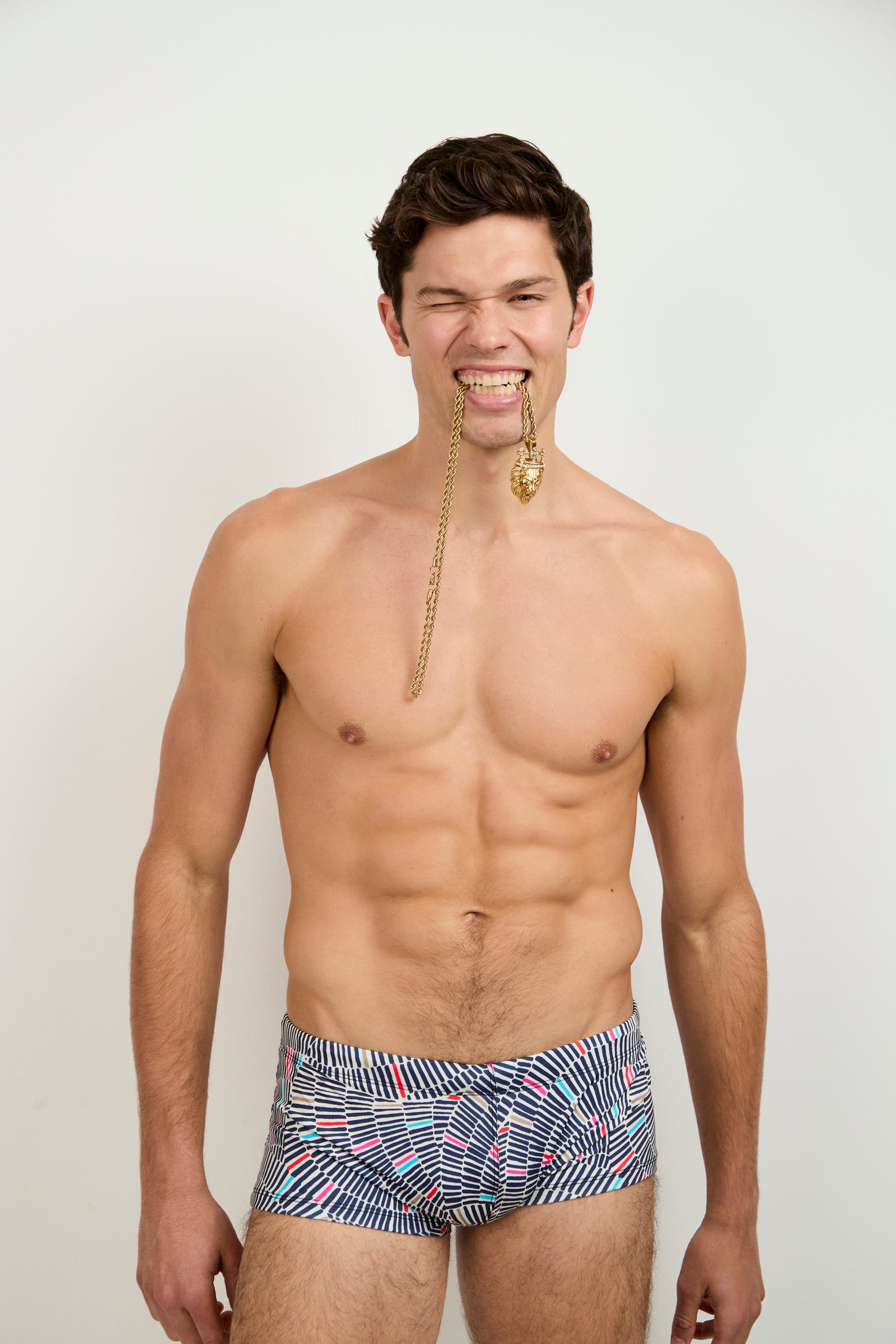 A shirtless man grins against a plain background, holding a long gold chain and pendant between his teeth while wearing the Sorrento Swim Trunk in Java.