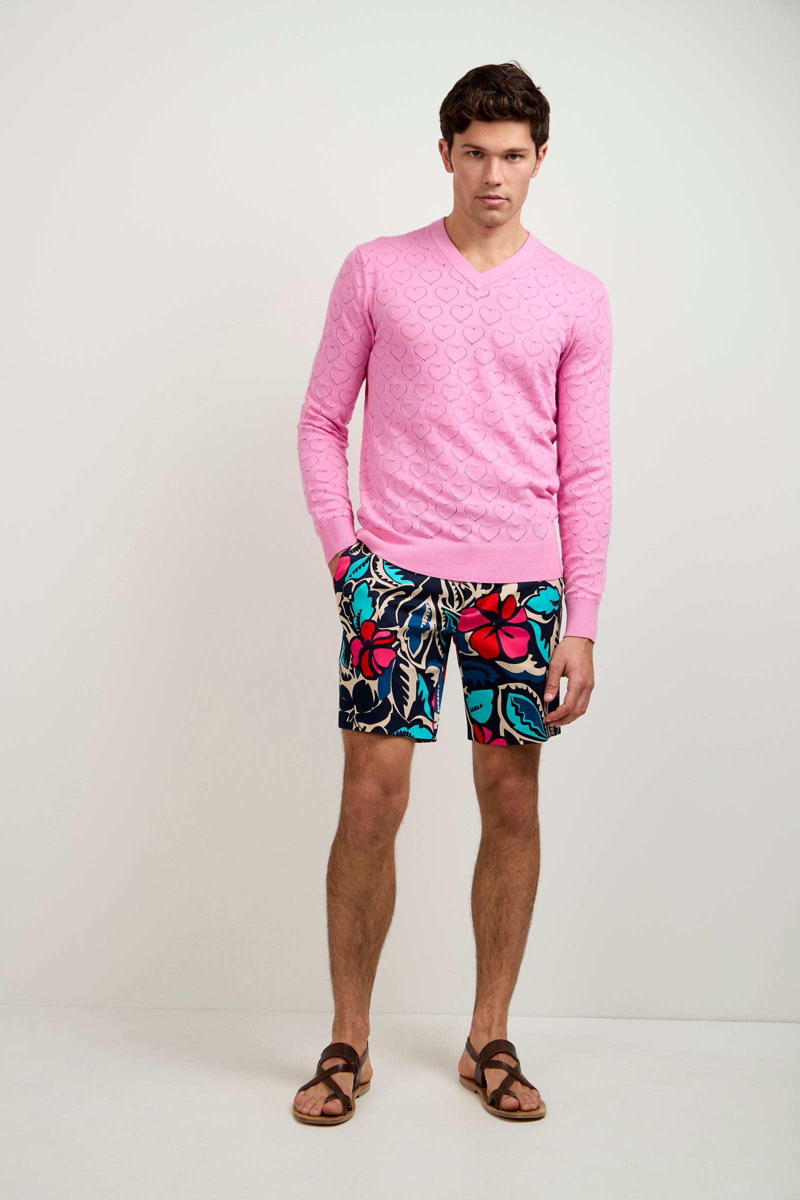 A man stands against a plain white wall wearing a light pink textured sweater, brown sandals, and the Lawrence Short - Copacabana with a colorful floral pattern. He has one hand in his pocket and maintains a neutral expression.