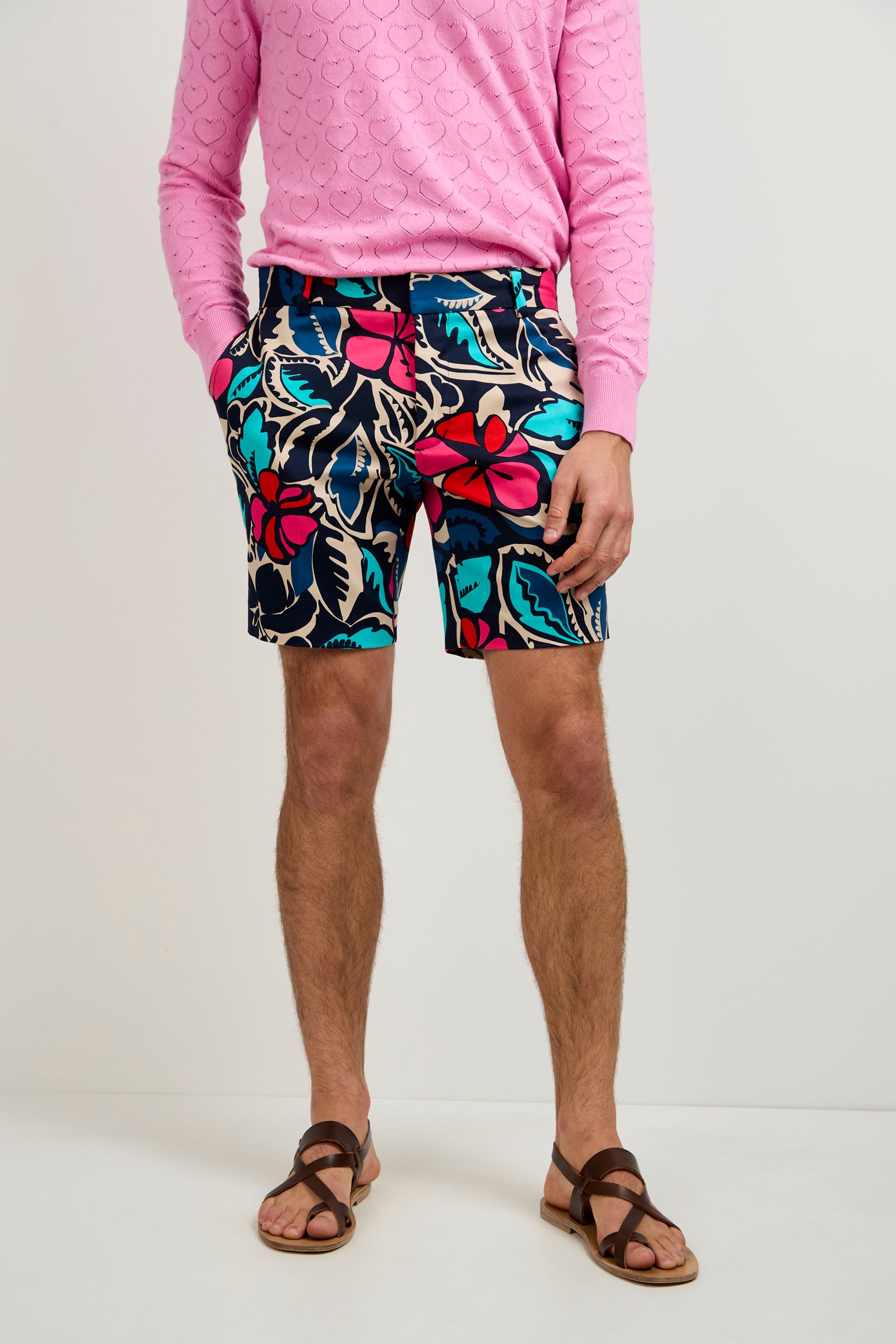 A person wears a pink textured sweater, Lawrence Short - Copacabana with bright floral patterns in red, blue, and teal, and brown sandals, standing against a plain light background.
