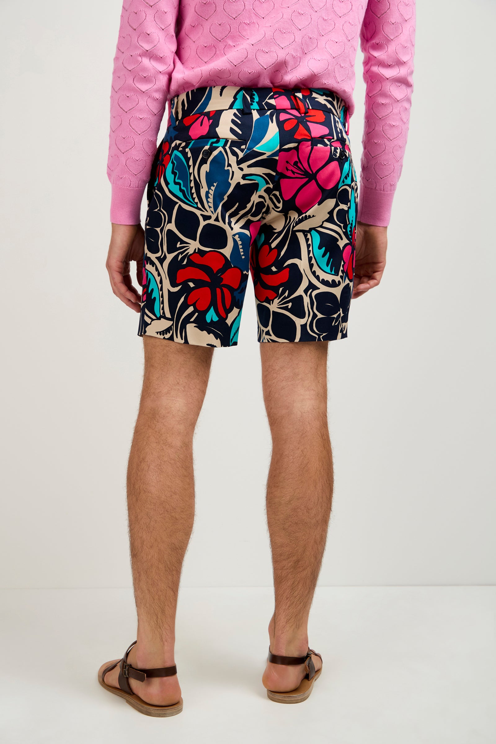 Wearing the Lawrence Short - Copacabana, featuring a colorful floral print in red, blue, and beige, a person stands with their back turned. Only their lower half is visible along with a pink textured sweater and brown sandals on a plain background.