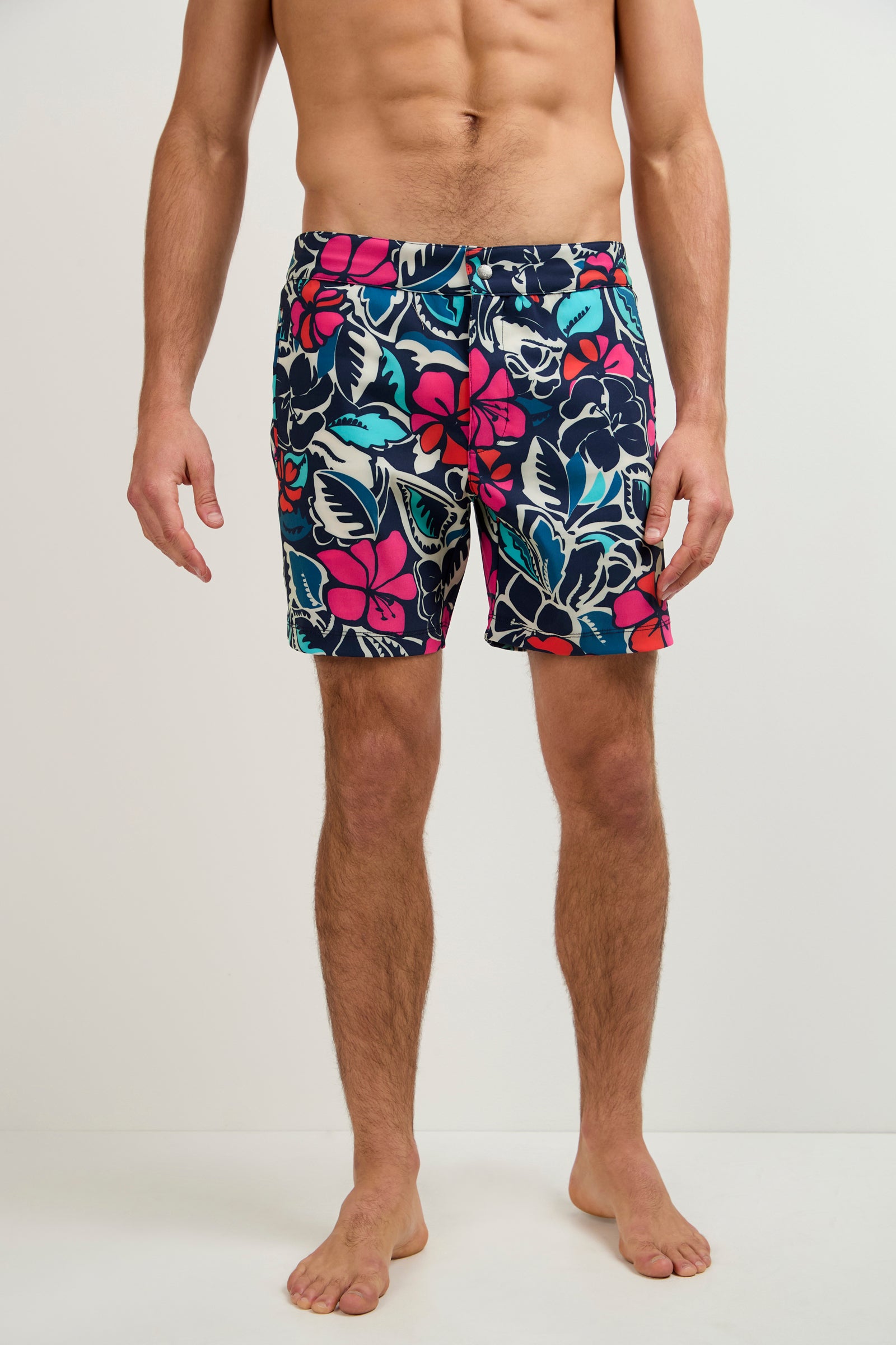 A man stands barefoot against a plain background, wearing the Honolulu Board Short - Copacabana, featuring vibrant pink, blue, and white tropical floral patterns. His upper body is bare with his hands resting at his sides.