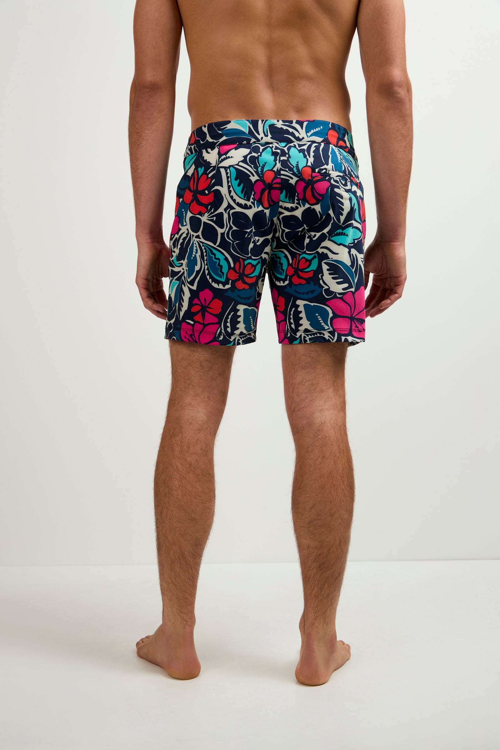 A man stands barefoot facing away, wearing the Honolulu Board Short - Copacabana with colorful floral patterns of red, pink, blue, and white against a plain background.