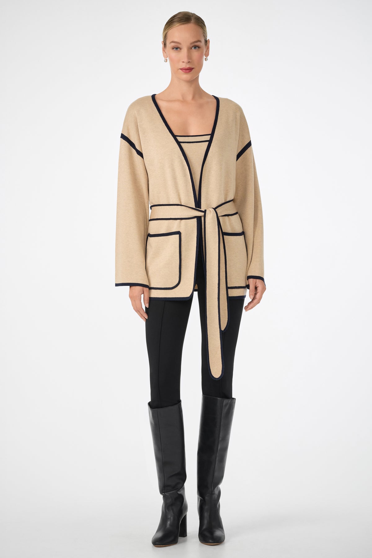 A woman stands facing forward in the Casady Knit Cami, paired with a beige cardigan featuring black trim and a waist tie, black pants, and knee-high black boots. Her hair is pulled back and her expression is neutral.