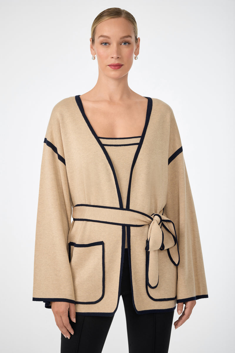 A woman wears the Wright Wrap Cardigan in beige with black trim, featuring wide sleeves, large front pockets, and a belted waist. She pairs it with a matching top and black pants, her hair pulled back, standing against a white background.