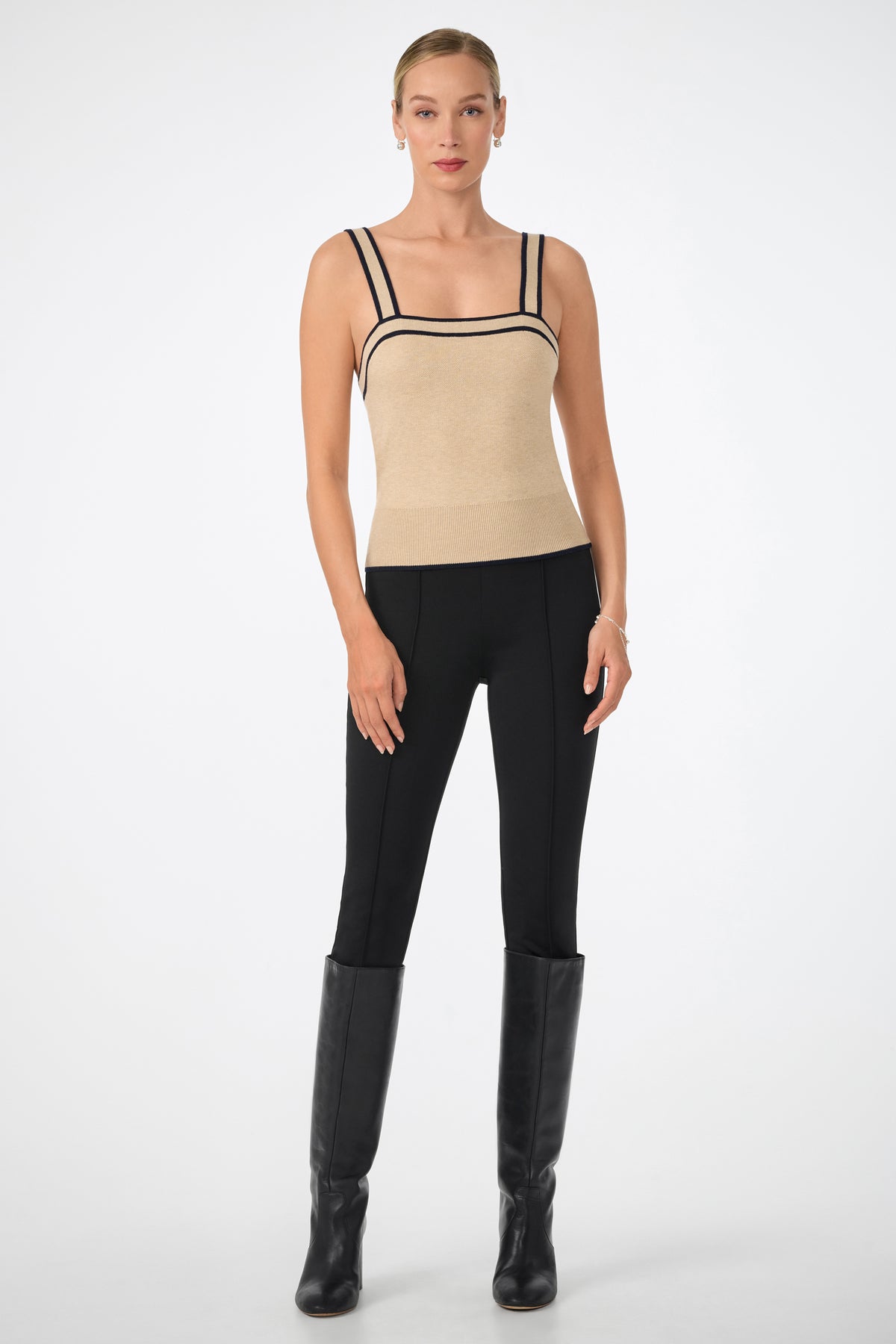 A woman stands against a plain background wearing the Casady Cami, a beige sleeveless top with black trim, paired with black fitted pants and knee-high boots. Her hair is pulled back and her arms rest relaxed at her sides.