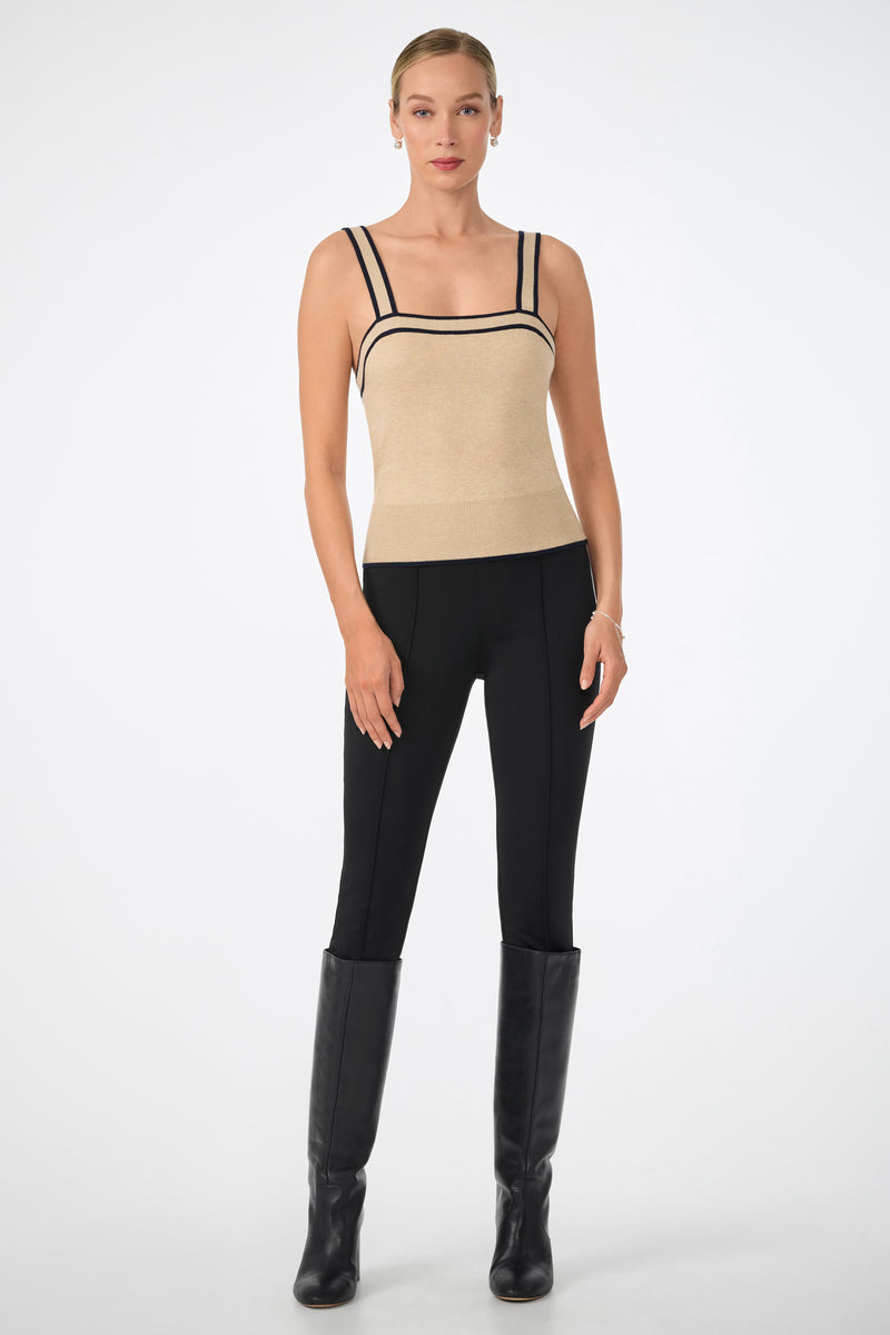 A woman stands against a plain background wearing the Casady Cami, a beige sleeveless top with black trim, paired with black fitted pants and knee-high boots. Her hair is pulled back and her arms rest relaxed at her sides.