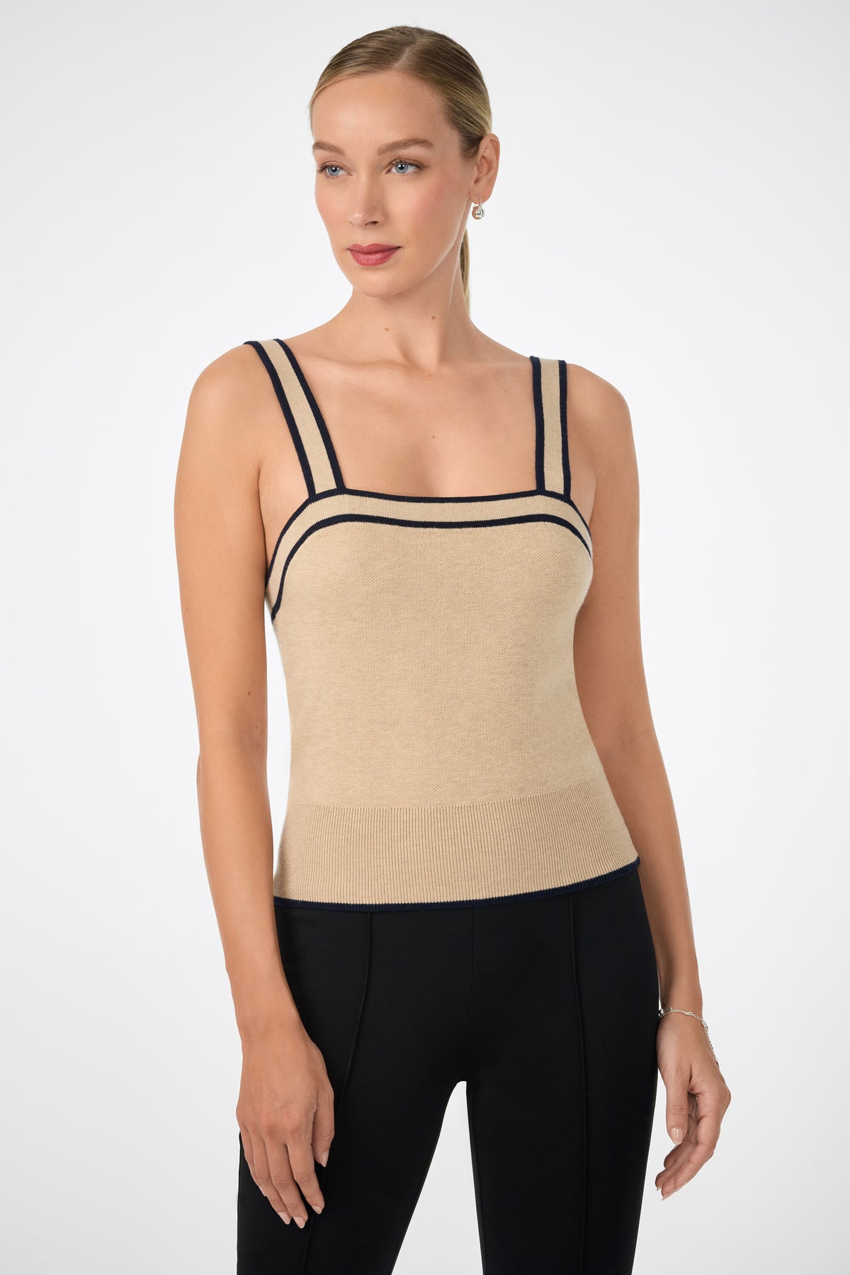 A woman wears the Casady Cami, a fitted beige tank top with black trim on the straps and neckline, paired with black pants, standing against a plain light background.
