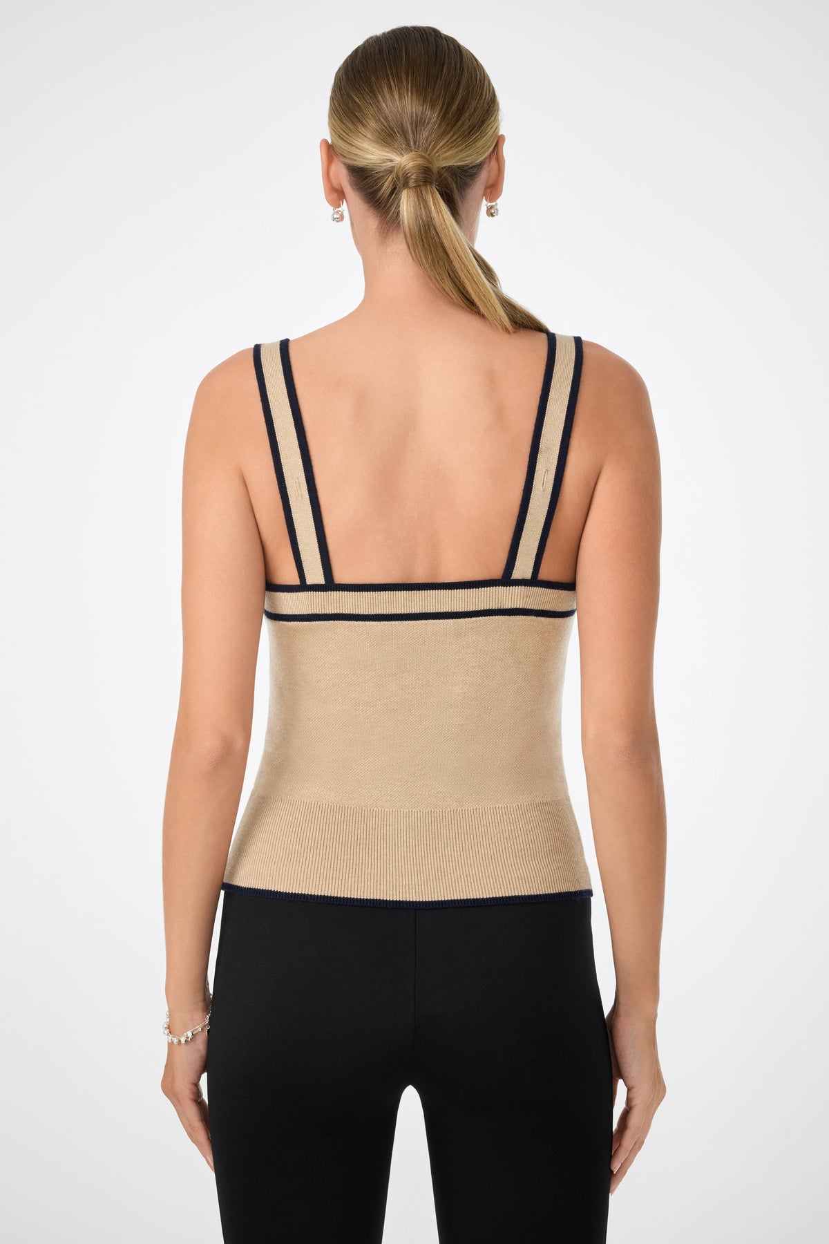 A woman with blonde hair in a low ponytail is shown from the back, wearing the Casady Cami—a beige tank top with black trim—and black fitted pants, standing against a plain light background.