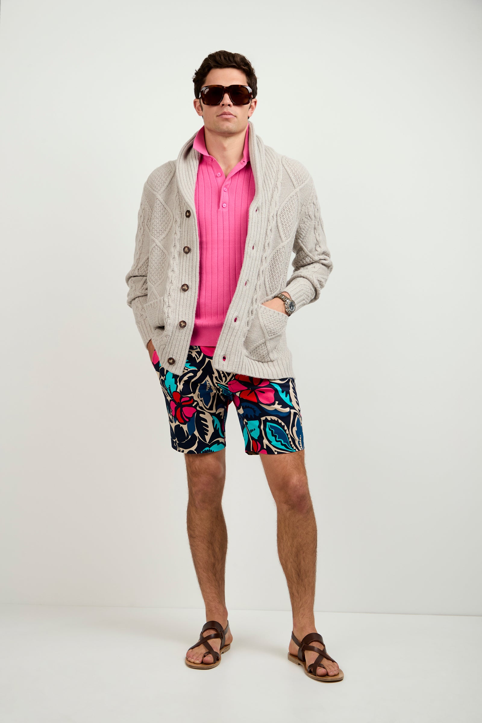 A man stands against a white background with his hands in his pockets, wearing sunglasses, the Brighton Cardigan (a beige knit cardigan) over a pink polo shirt, colorful floral shorts, and brown sandals.