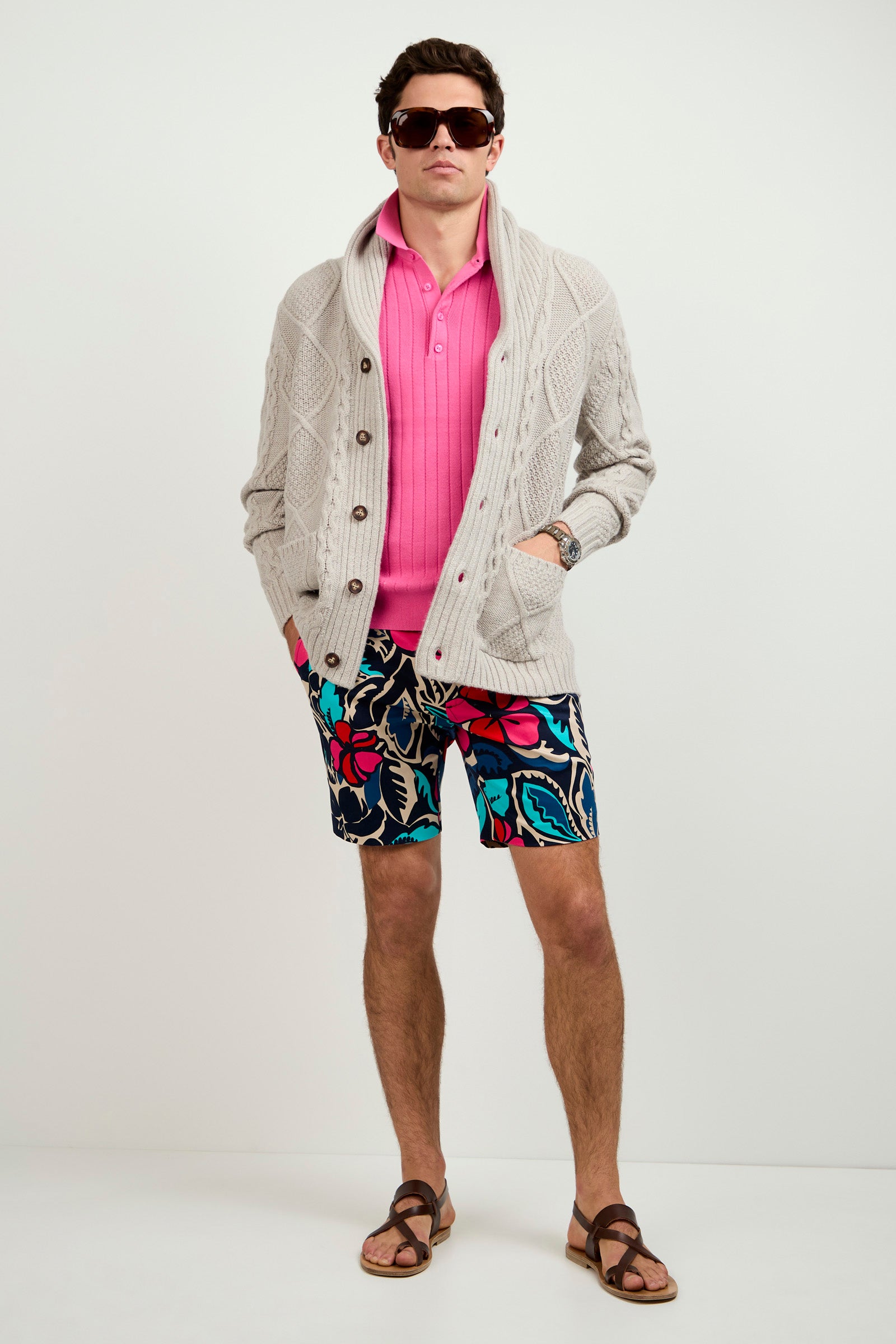A man stands against a plain background wearing sunglasses, a pink polo, the Brighton Cardigan, colorful patterned shorts, and brown sandals with his hands in his pockets.
