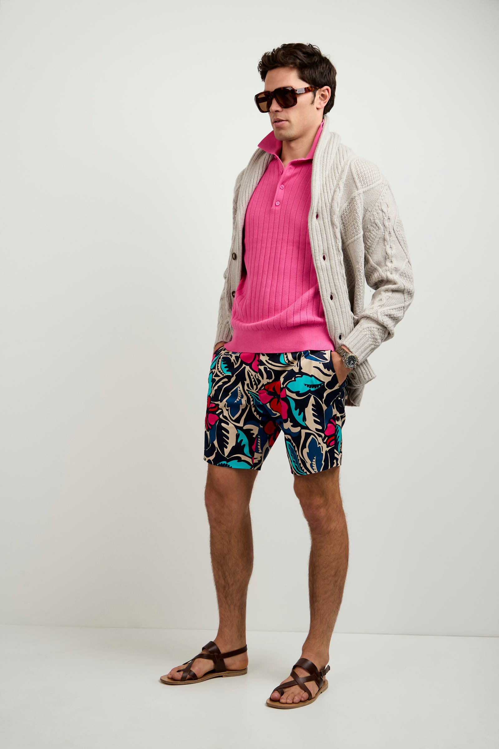A man stands against a plain white background wearing the Brighton Cardigan over a bright pink shirt, colorful patterned shorts, brown sandals, and large sunglasses.