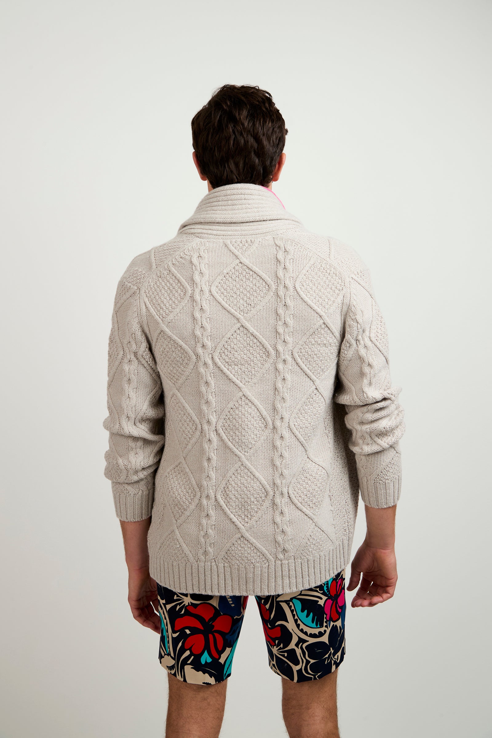 A man faces away, wearing the Brighton Cardigan—a cream cable-knit sweater with a shawl collar—and colorful floral shorts, standing against a plain light background.