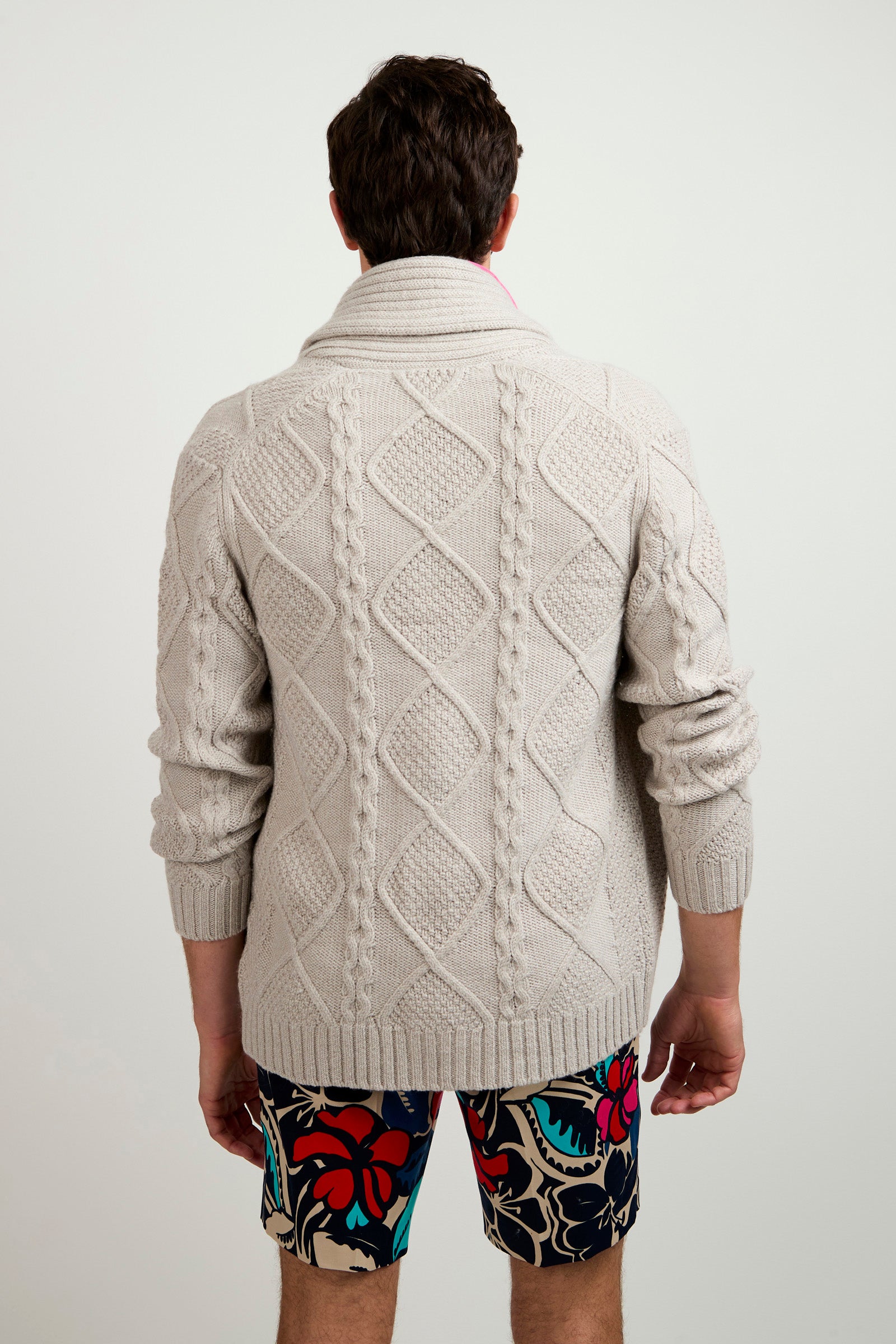 A man stands facing away, wearing the Brighton Cardigan—a light gray, patterned knit sweater—and colorful floral shorts against a plain white background.