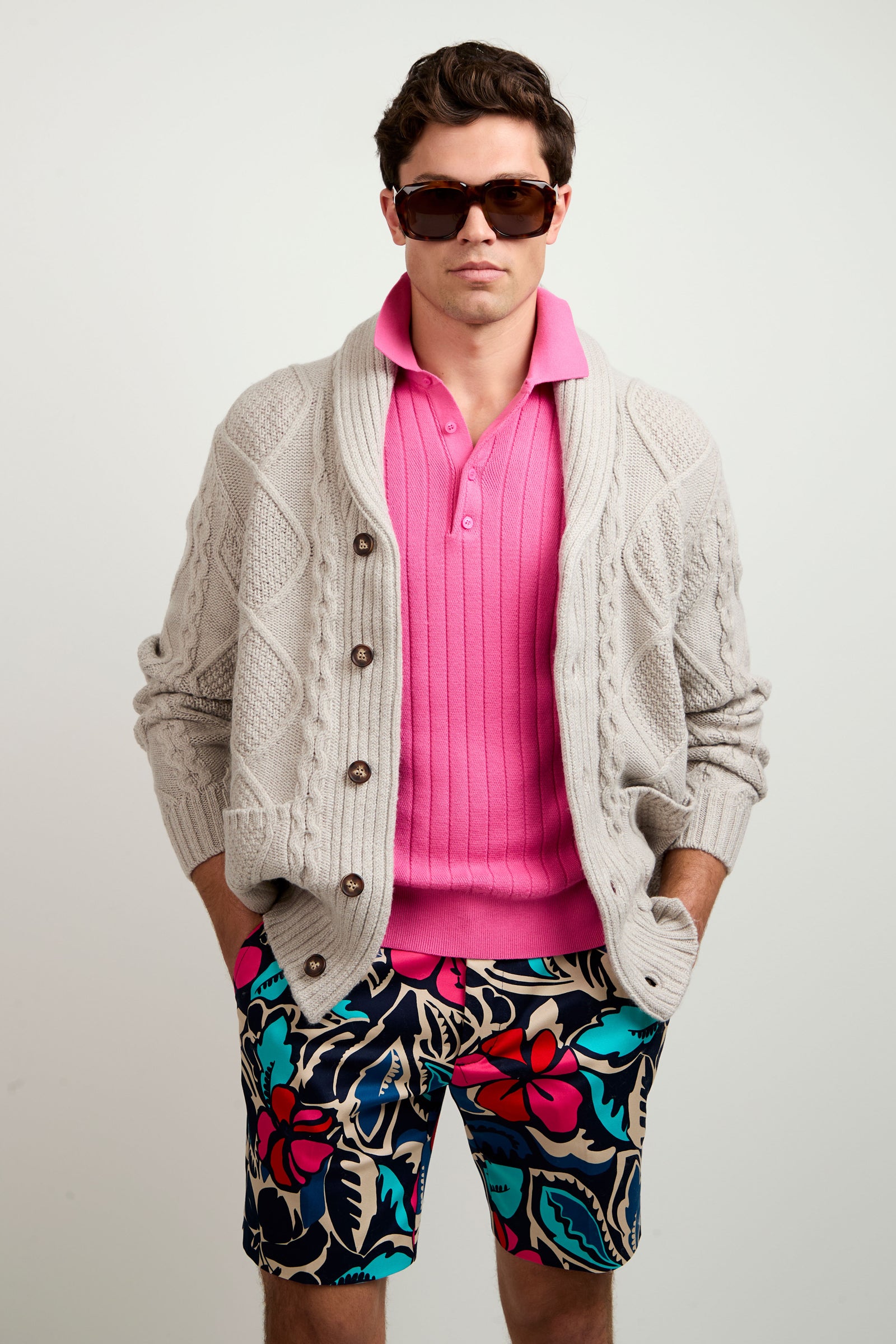 A man stands with hands in pockets against a plain background, wearing the Brighton Cardigan in light gray over a bright pink polo shirt, paired with large sunglasses and colorful floral shorts.