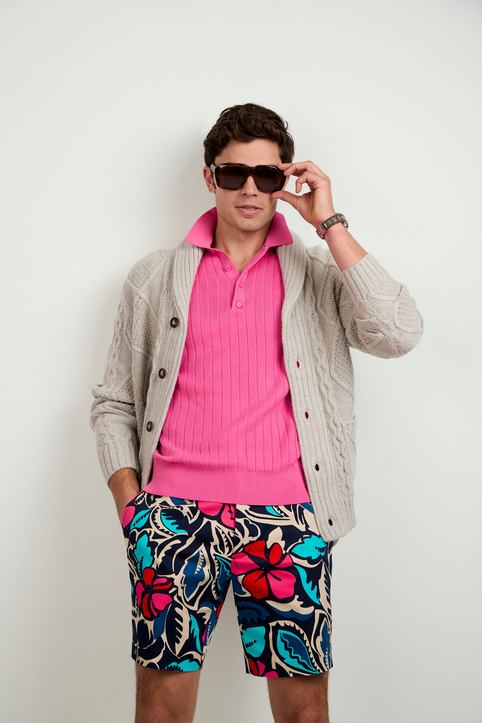 A man stands against a plain white background wearing sunglasses, the Brighton Cardigan in beige, a bright pink polo shirt, colorful floral shorts, and has one hand in his pocket.