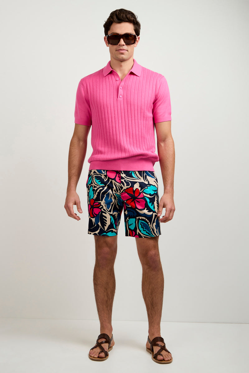 A man in sunglasses, the Alvarez Short-Sleeve Polo in bright pink, colorful floral shorts, and brown sandals stands against a plain white background.