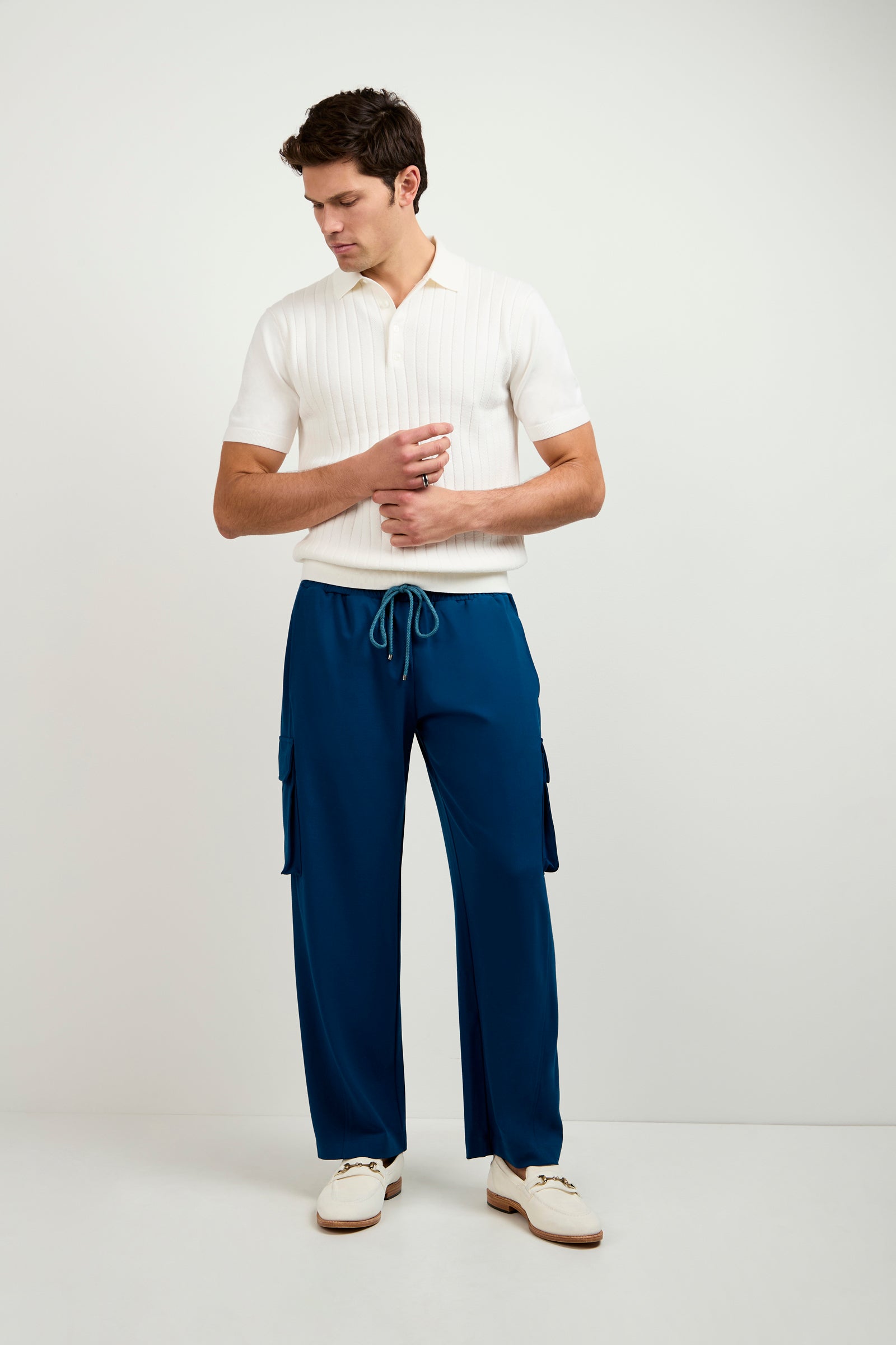 A man stands against a plain white background wearing the Alvarez Short-Sleeve Polo in white, paired with blue drawstring cargo pants and white shoes. He looks down with his hands resting on his pants.