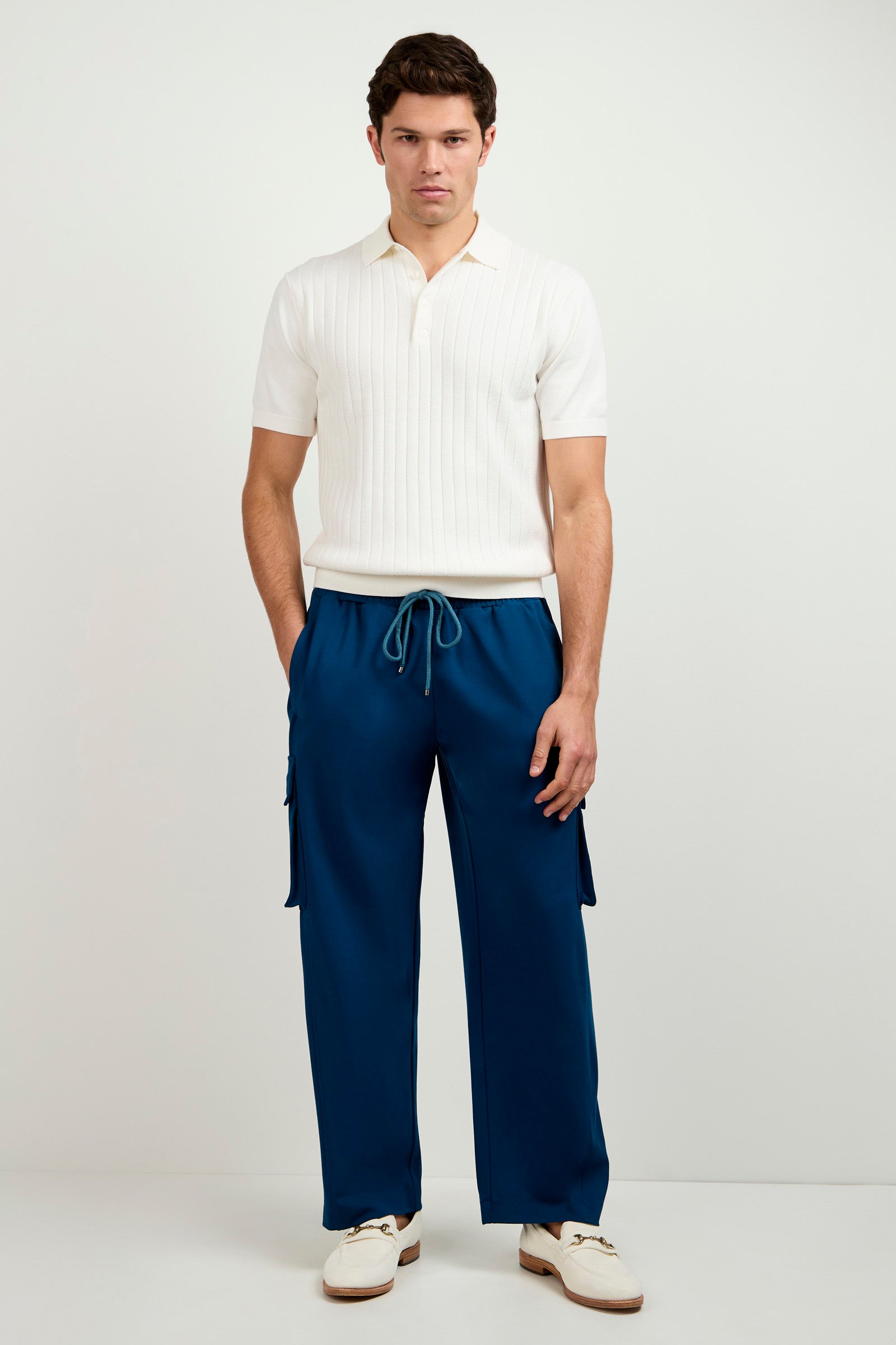 A man stands against a light background, wearing a short-sleeved white knit polo shirt, Lounge Cargo Pant in blue with side pockets and drawstring waist, and white loafers. His hands are in his pockets as he looks straight ahead.