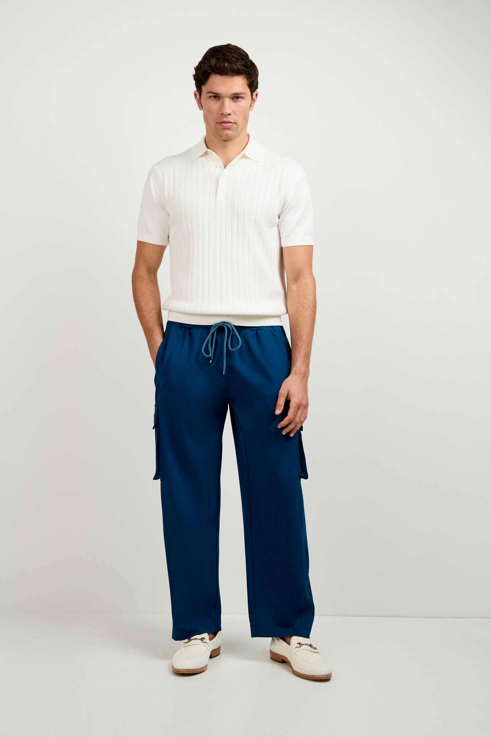 A man with a neutral expression stands against a white background wearing blue Lounge Cargo Pants with side pockets, a short-sleeve white knit polo, and white sneakers. His hands are in his pockets.