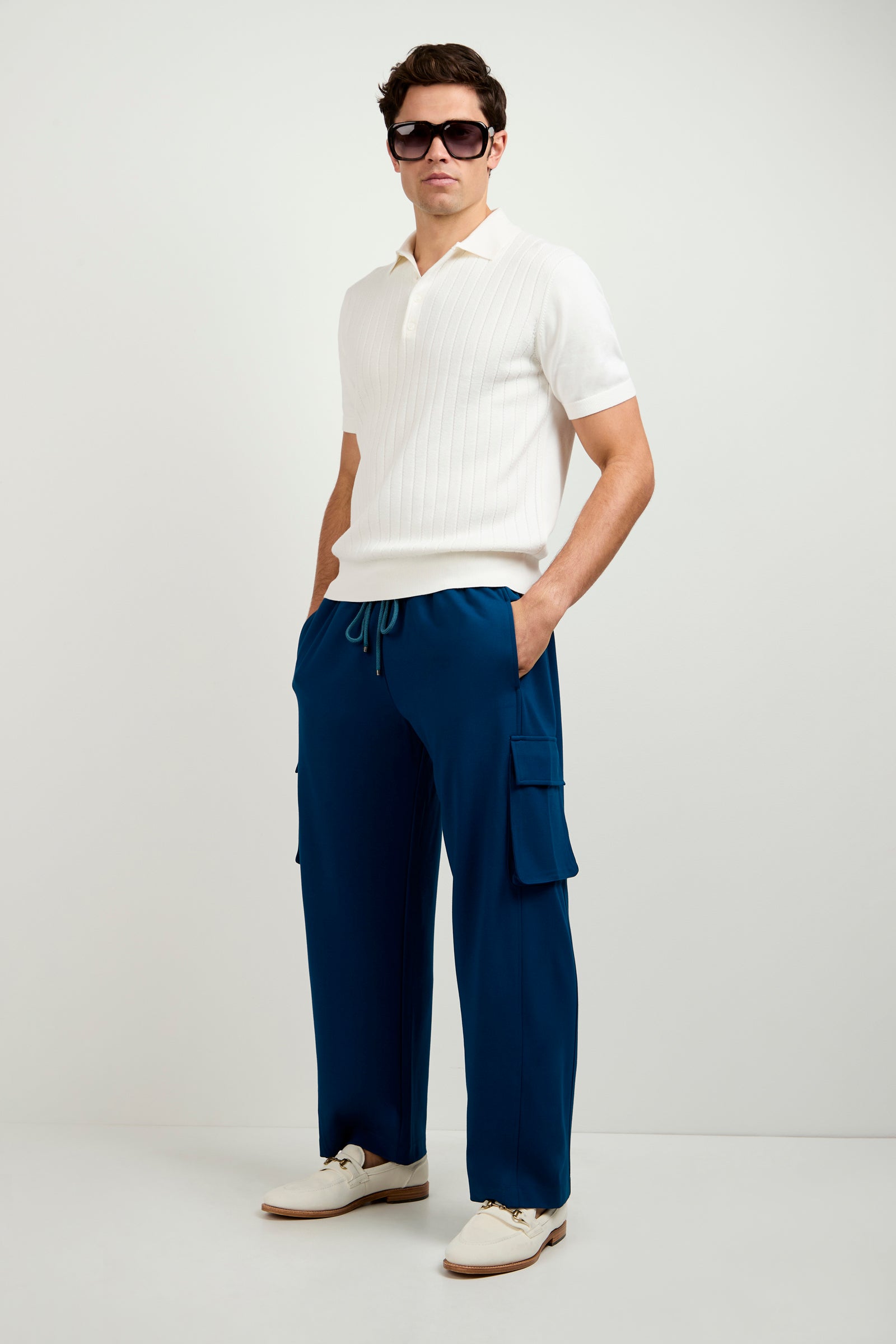 A man wearing Lounge Cargo Pant in blue, a white short-sleeve knit polo, white shoes, and sunglasses stands with hands in pockets against a plain white background.