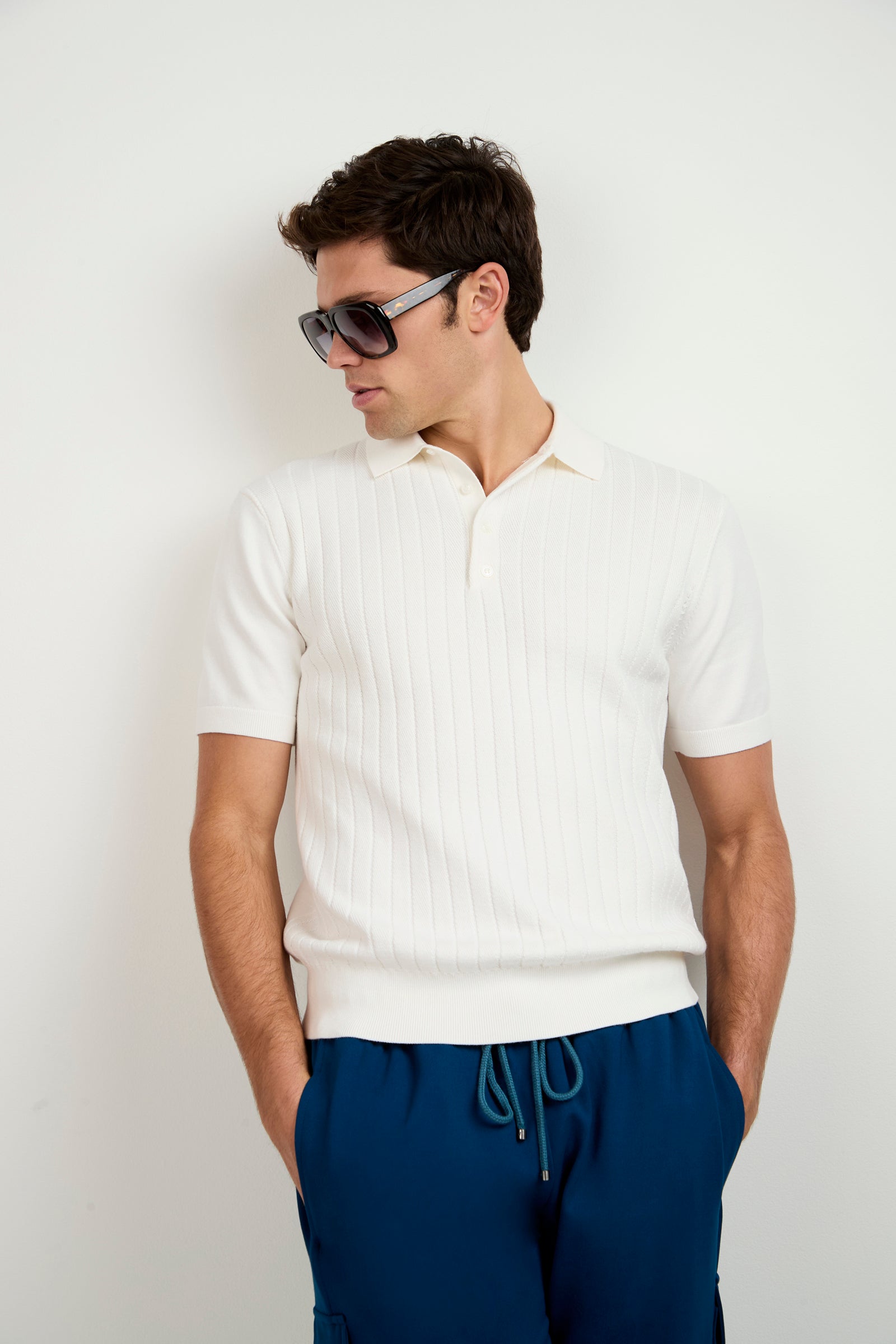 A man dressed in the Alvarez Short-Sleeve Polo and blue drawstring pants, wearing sunglasses, stands with his hands in his pockets against a plain white background, looking to his left.