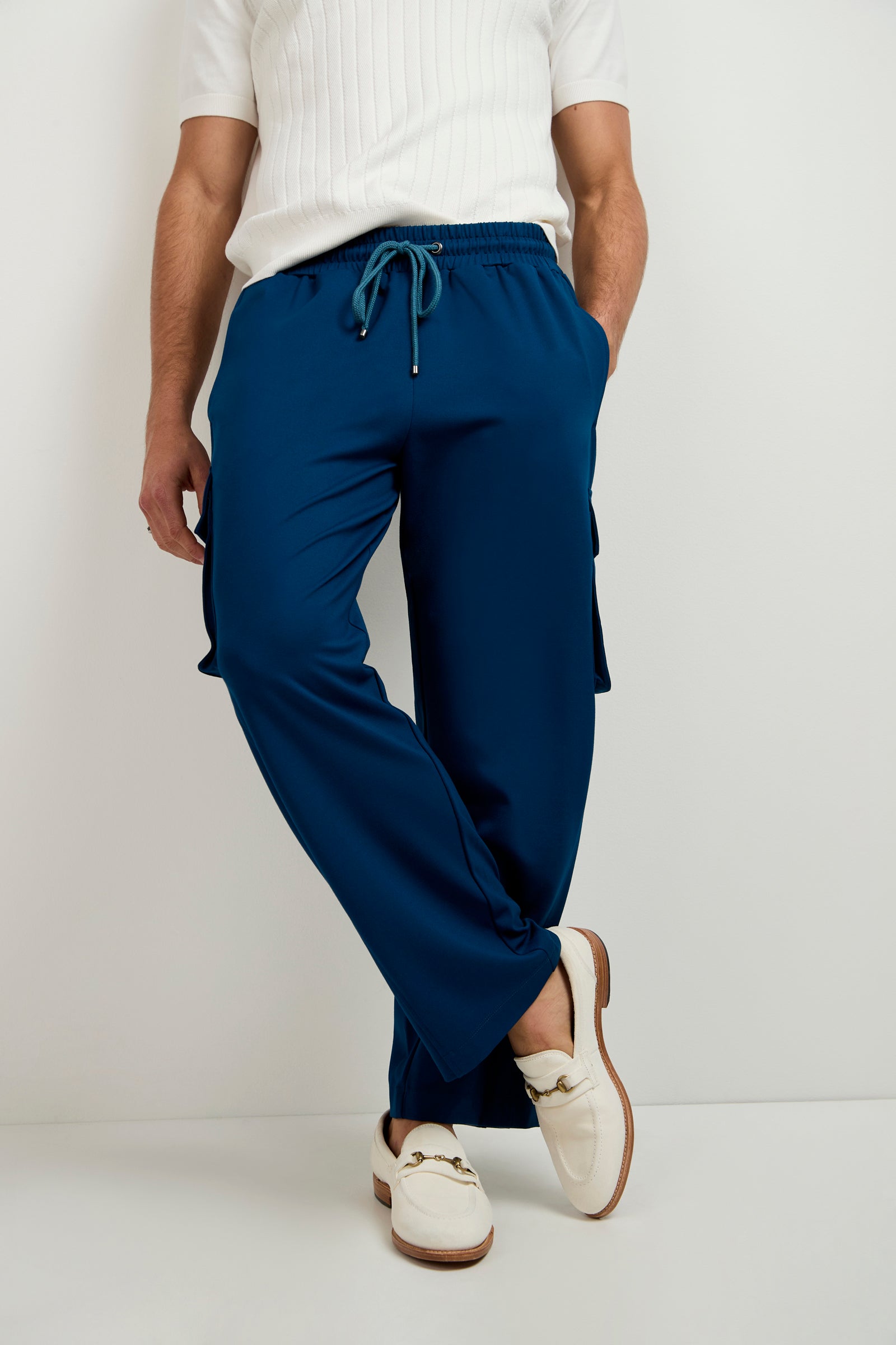 A person stands against a white background wearing a white short-sleeve ribbed shirt, Lounge Cargo Pant in blue with pockets and drawstring, and white loafers featuring gold hardware.