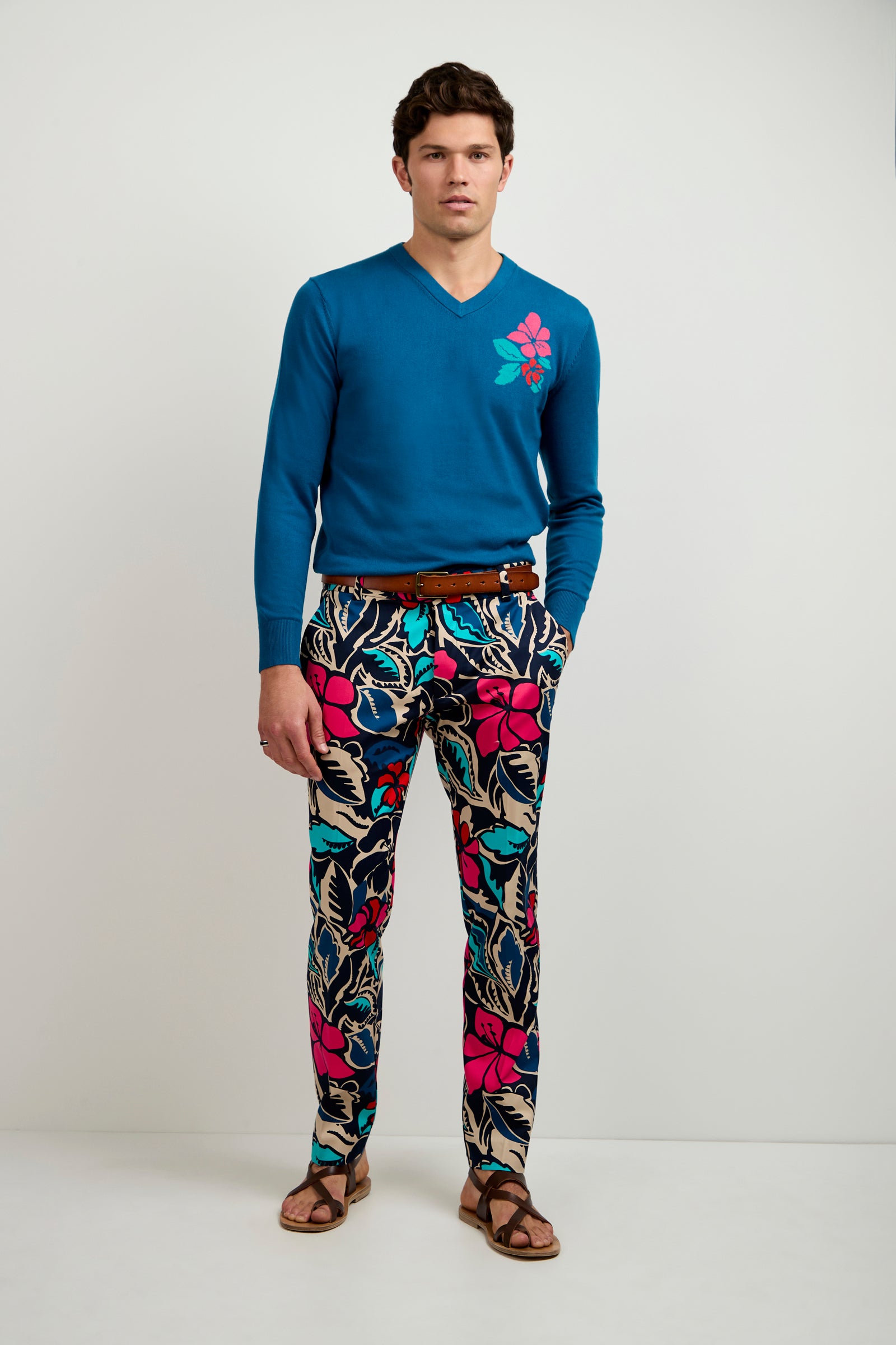 A man stands against a plain background wearing a teal sweater with a pink flower design, Clyde Slim Trouser - Copacabana featuring colorful blue, black, and pink floral patterns, a brown belt, and brown sandals.