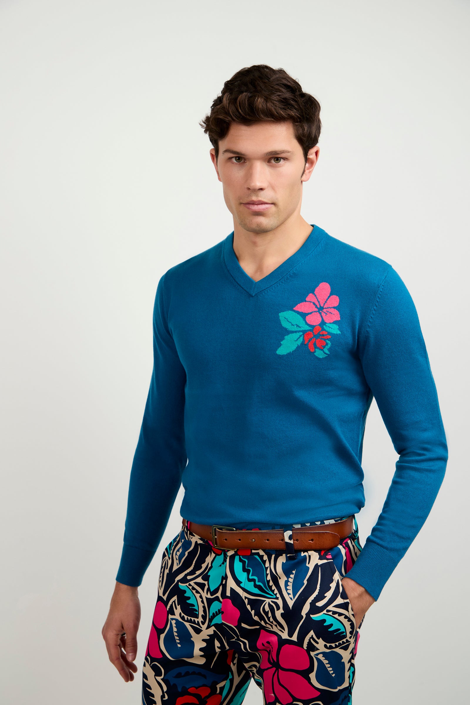 A young man with short brown hair wears the Copacabana V-Neck Sweater, featuring a pink and teal floral chest design, paired with colorful tropical-patterned pants and a brown belt, standing against a plain light background.