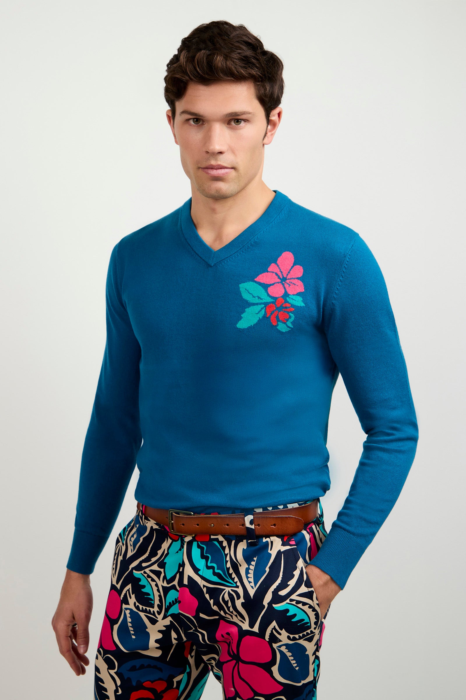 A man wearing the Copacabana V-Neck Sweater with a pink and teal floral chest design, paired with colorful abstract floral and leaf patterned pants, stands against a plain light background.