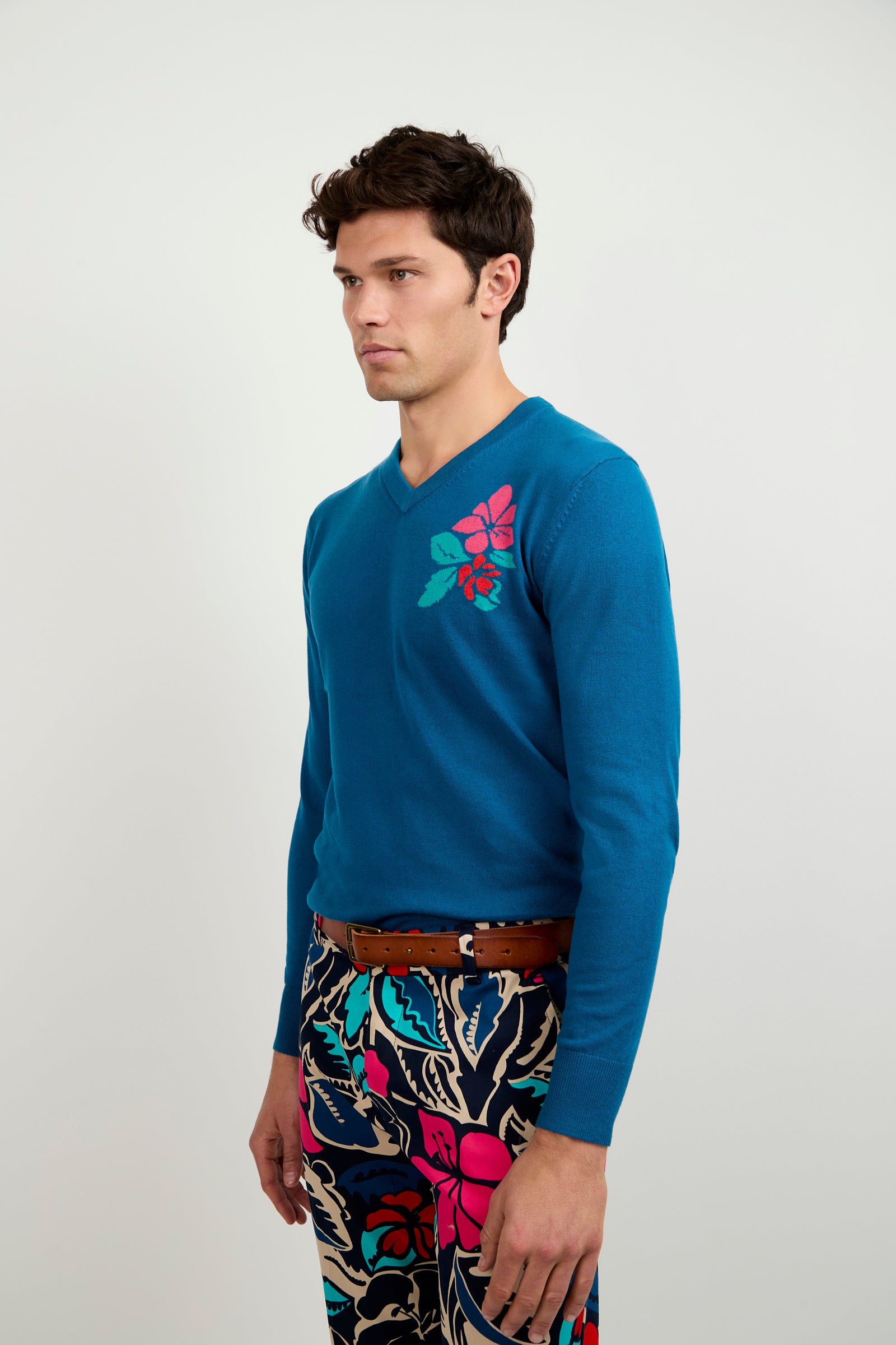 A man with short dark hair wears the Copacabana V-Neck Sweater, featuring a bright blue color and a pink and green floral shoulder design, paired with bold floral patterned pants. He stands sideways against a plain light background.