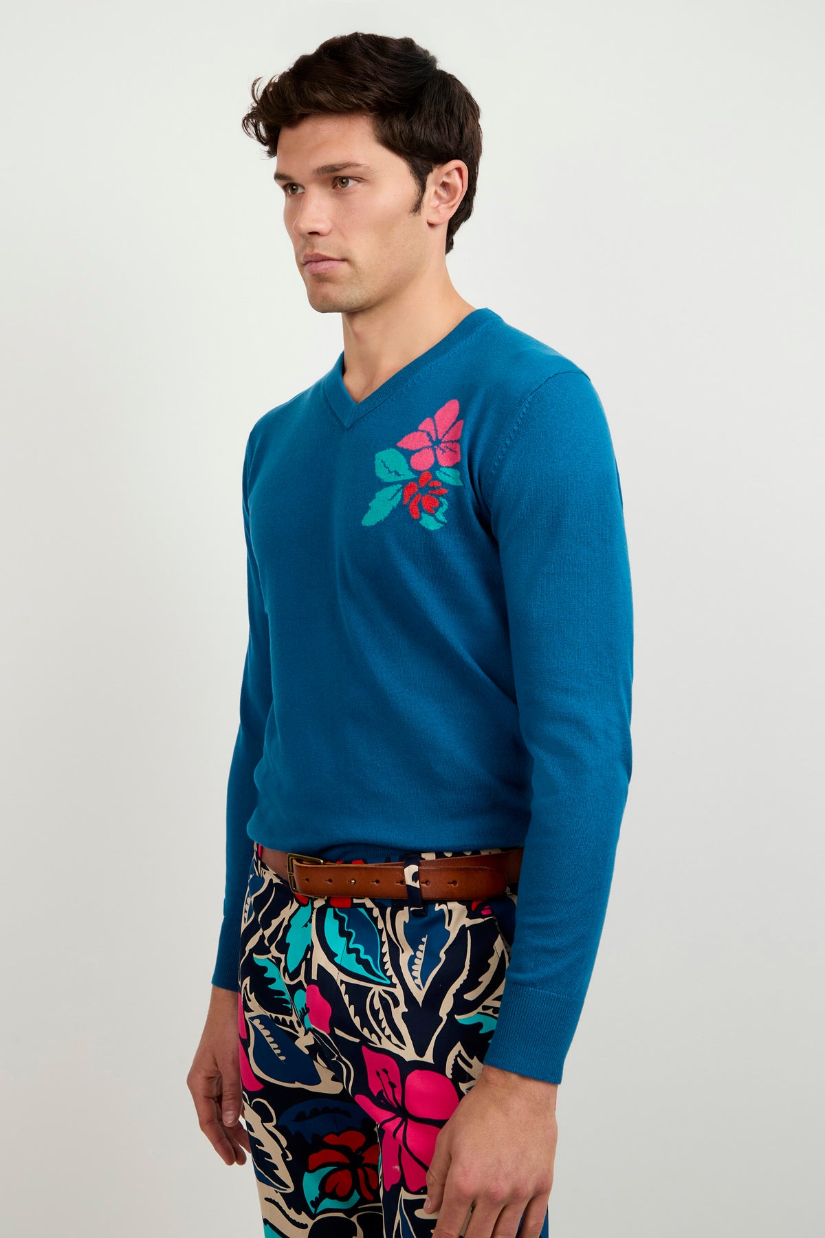 A man in profile wears the Copacabana V-Neck Sweater with pink and red floral embroidery on the shoulder, paired with navy pants patterned with bold, colorful leaves and flowers.
