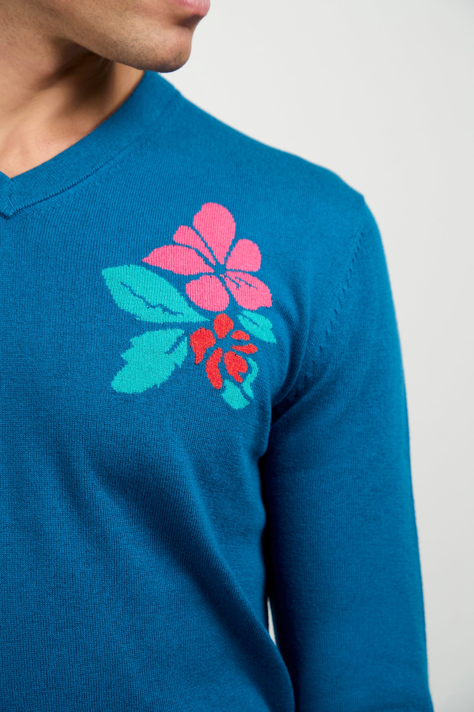 A person wearing the Copacabana V-Neck Sweater—a blue top with a pink and red floral design with green leaves on the upper left chest. Only their lower face, neck, and upper torso are visible.