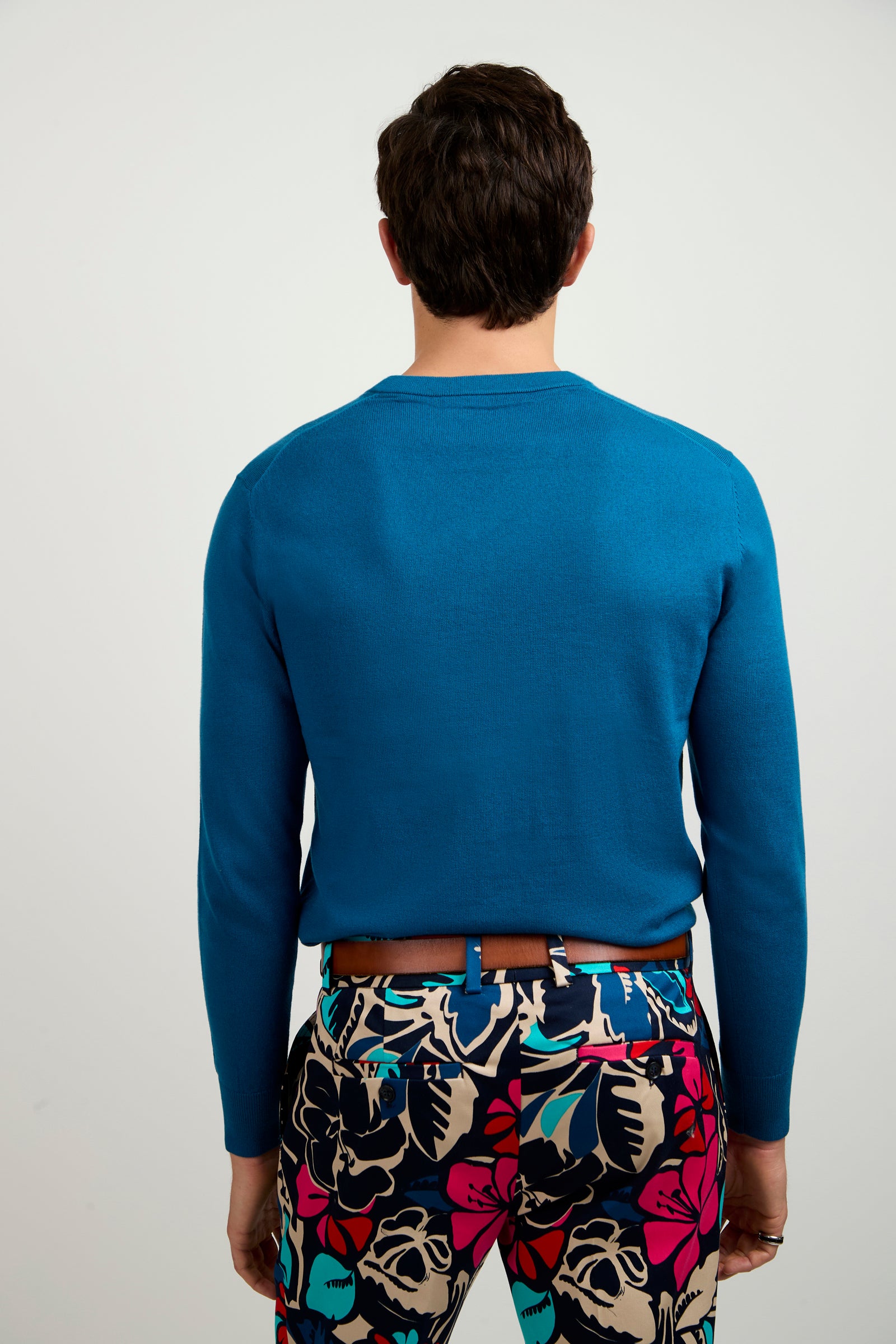 A person stands facing away, wearing the Copacabana V-Neck Sweater in bright blue and colorful floral-patterned pants with pink, blue, and beige on a plain light background.