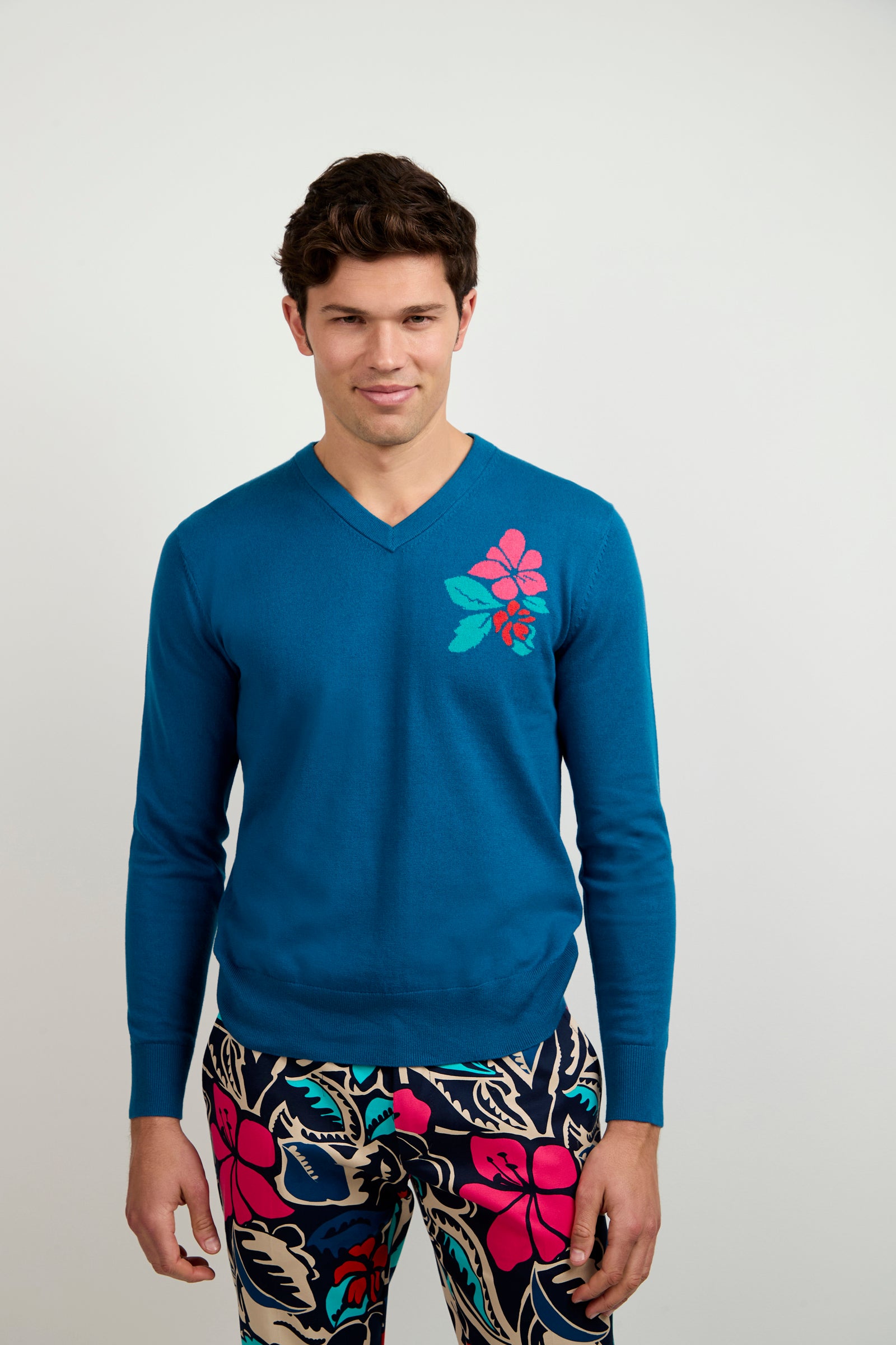 A man wears the Copacabana V-Neck Sweater, featuring a blue base with a pink and green flower design on the chest, and colorful floral-patterned pants, standing against a plain light background.
