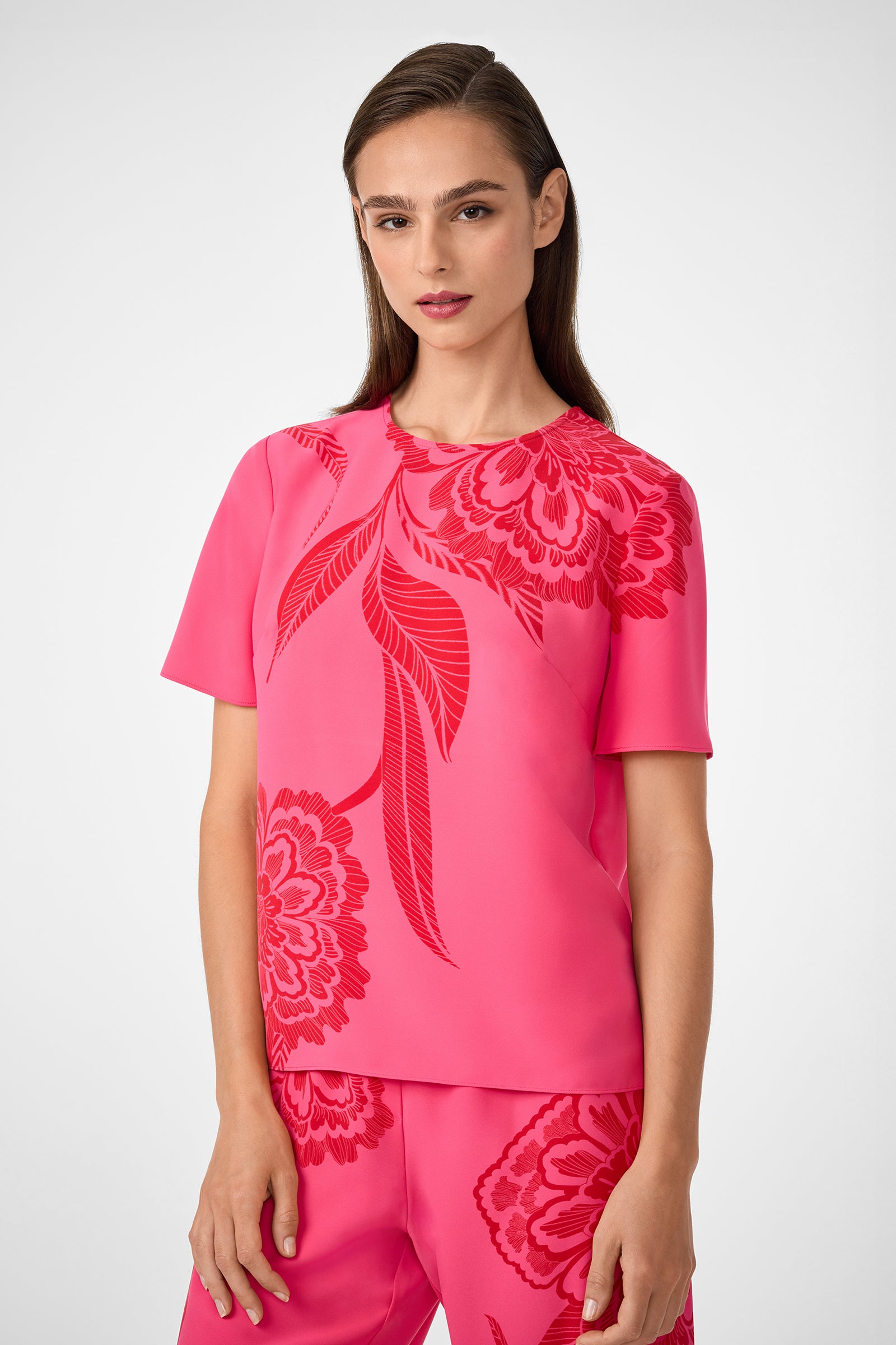 A woman with straight brown hair wears the Sugarhill Top, a bright pink top with large red floral patterns. She stands against a light gray background, looking directly at the camera with a neutral expression.