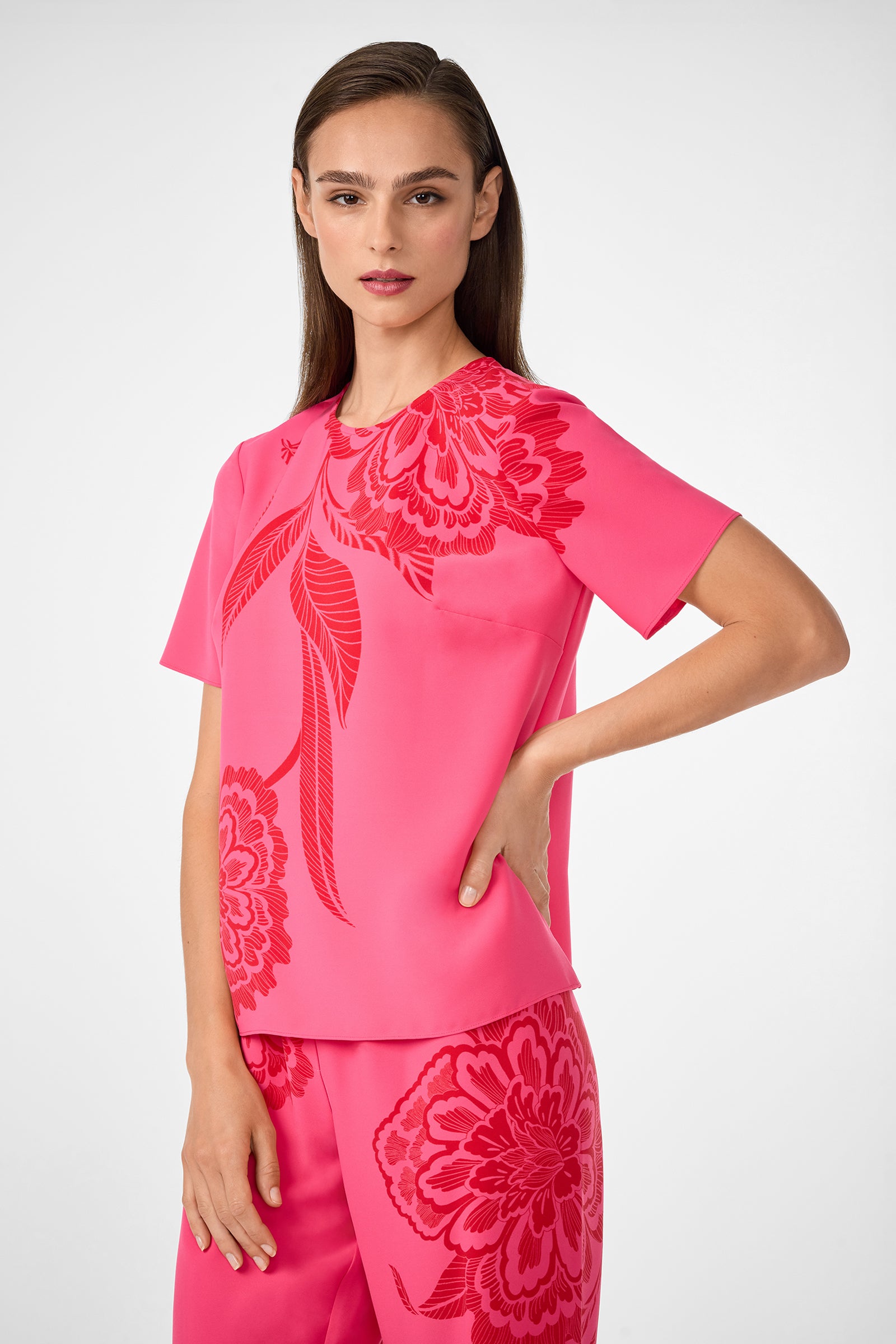 A woman with straight brown hair wears the Sugarhill Top, a bright pink short-sleeve top featuring bold red floral patterns. She stands against a light background with one arm bent at her waist.