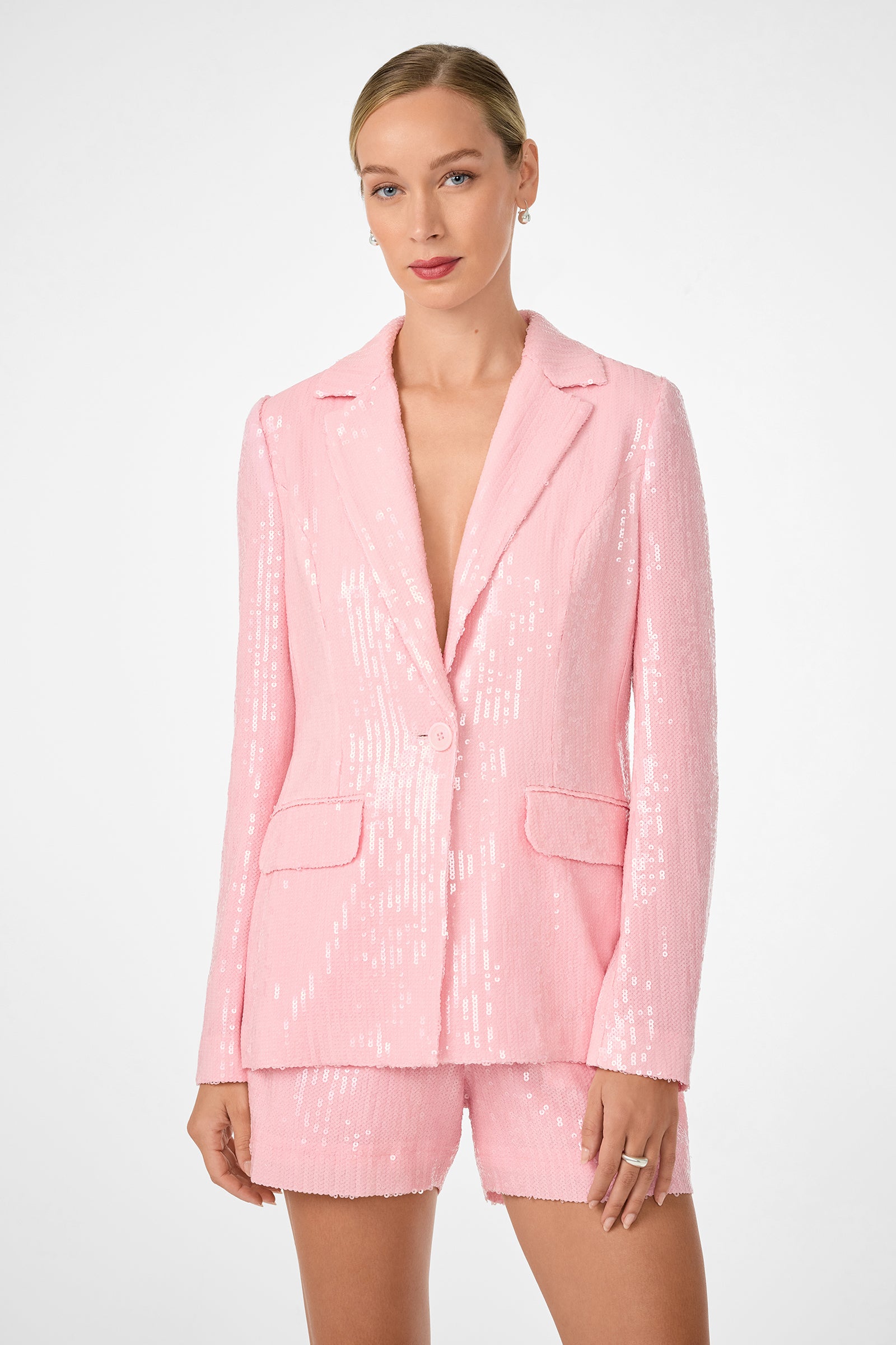 A woman models the Alternates 2 Blazer in light pink sequins with matching shorts against a plain white background. Her hair is pulled back, and she looks directly at the camera with a subtle smile.