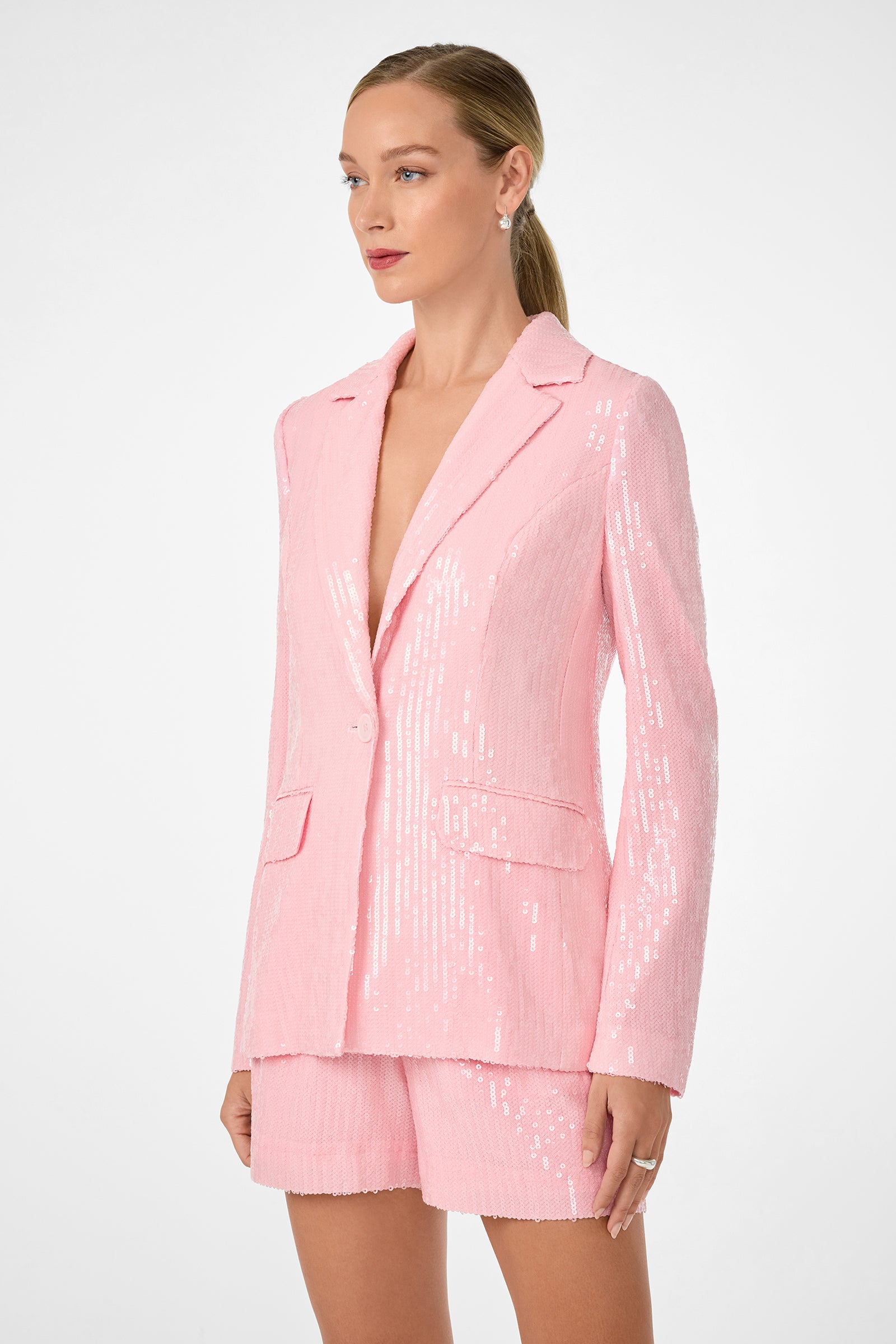 A woman with blonde hair in a low ponytail wears the light pink, sequined Alternates 2 Blazer and matching shorts, standing against a plain white background and looking slightly to the side.