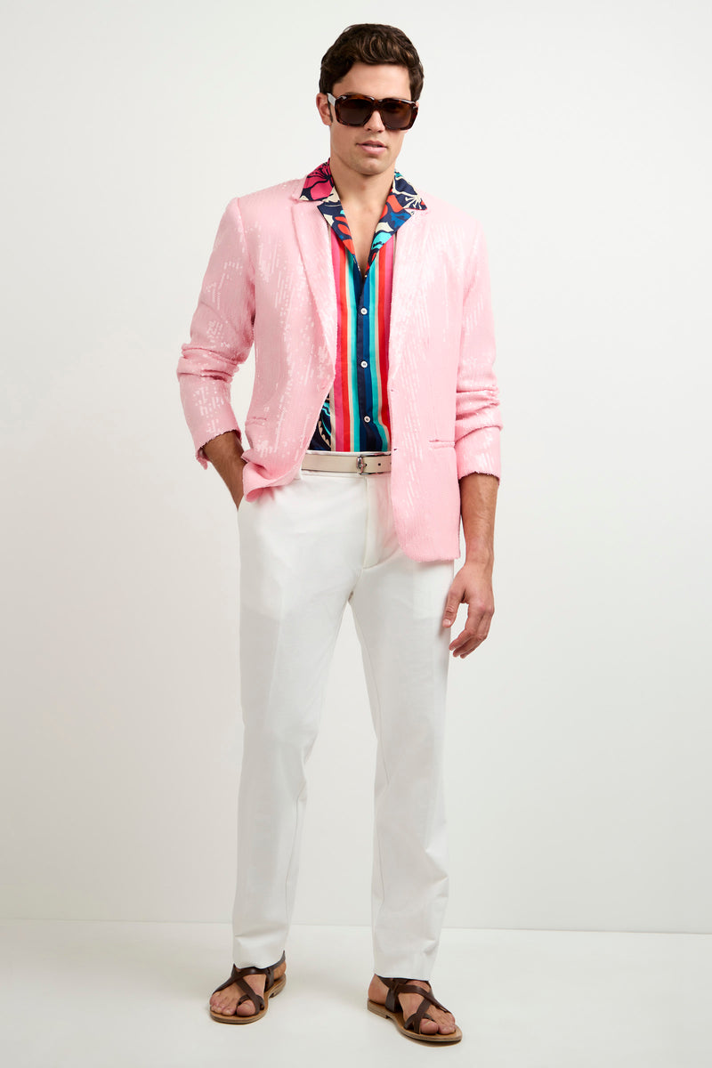 A man stands confidently against a white background wearing the Bowie Sequin Blazer, a colorful patterned shirt, white pants, dark sunglasses, and brown sandals. His left hand is in his pocket.