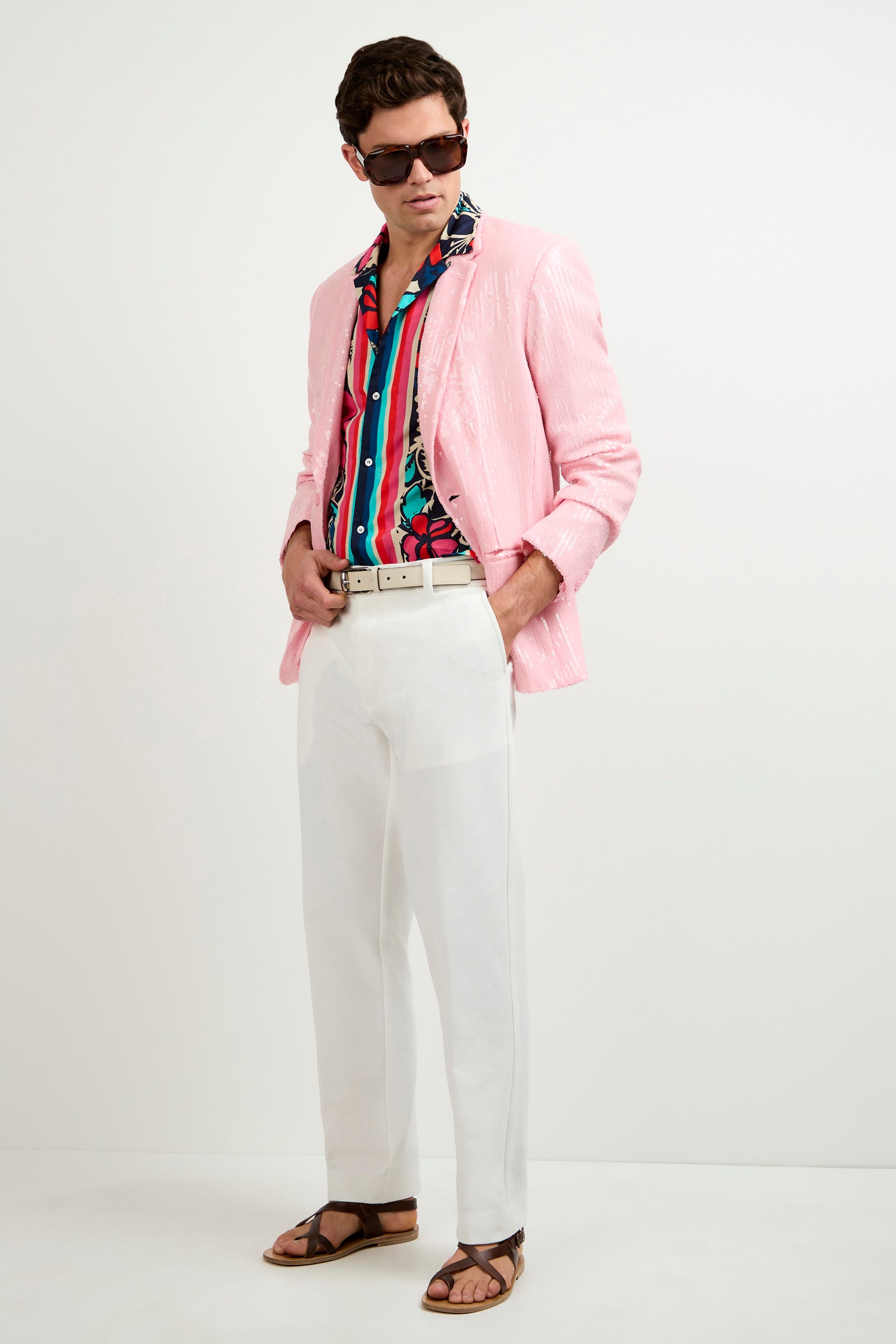 A man stands with hands in his pockets against a plain background, wearing the Bowie Sequin Blazer, a colorful floral shirt, white trousers, a white belt, brown sandals, and large sunglasses while looking to the side.