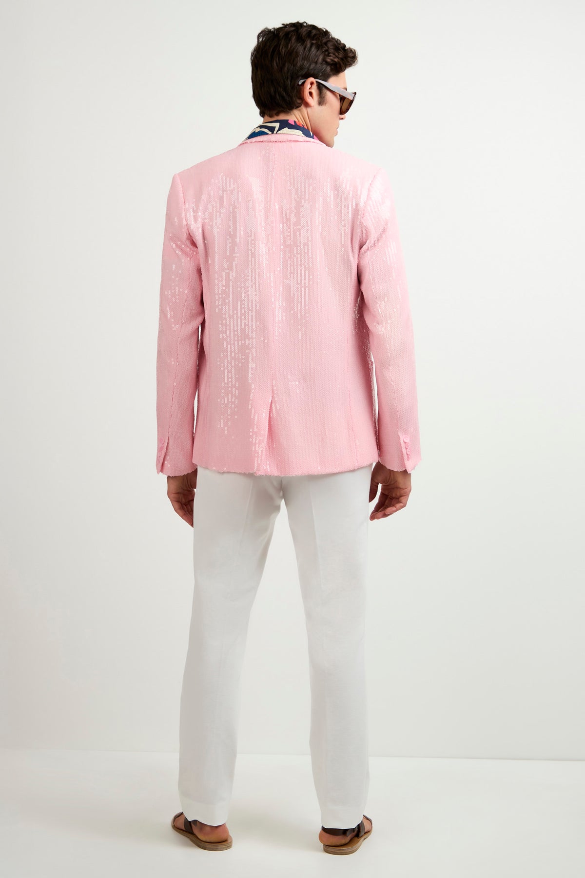 A man stands with his back to the camera, wearing the Bowie Sequin Blazer in shiny pink, paired with white pants, brown sandals, sunglasses, and a colorful scarf. The background is plain white.