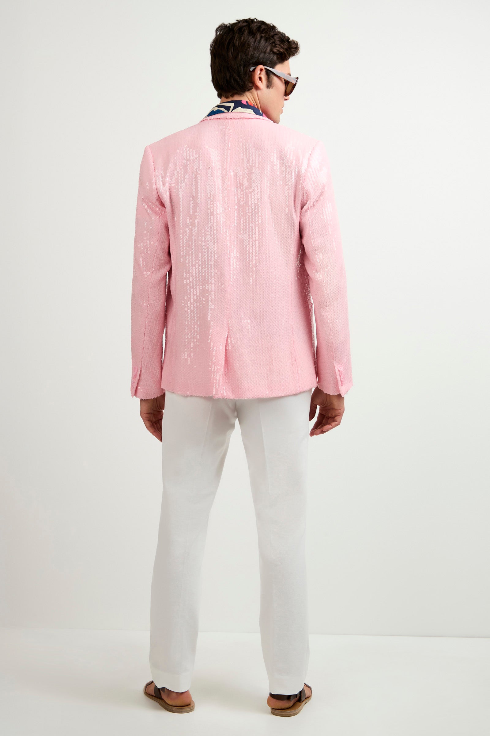A man stands with his back to the camera, wearing the Bowie Sequin Blazer in shiny pink, paired with white pants, brown sandals, sunglasses, and a colorful scarf. The background is plain white.