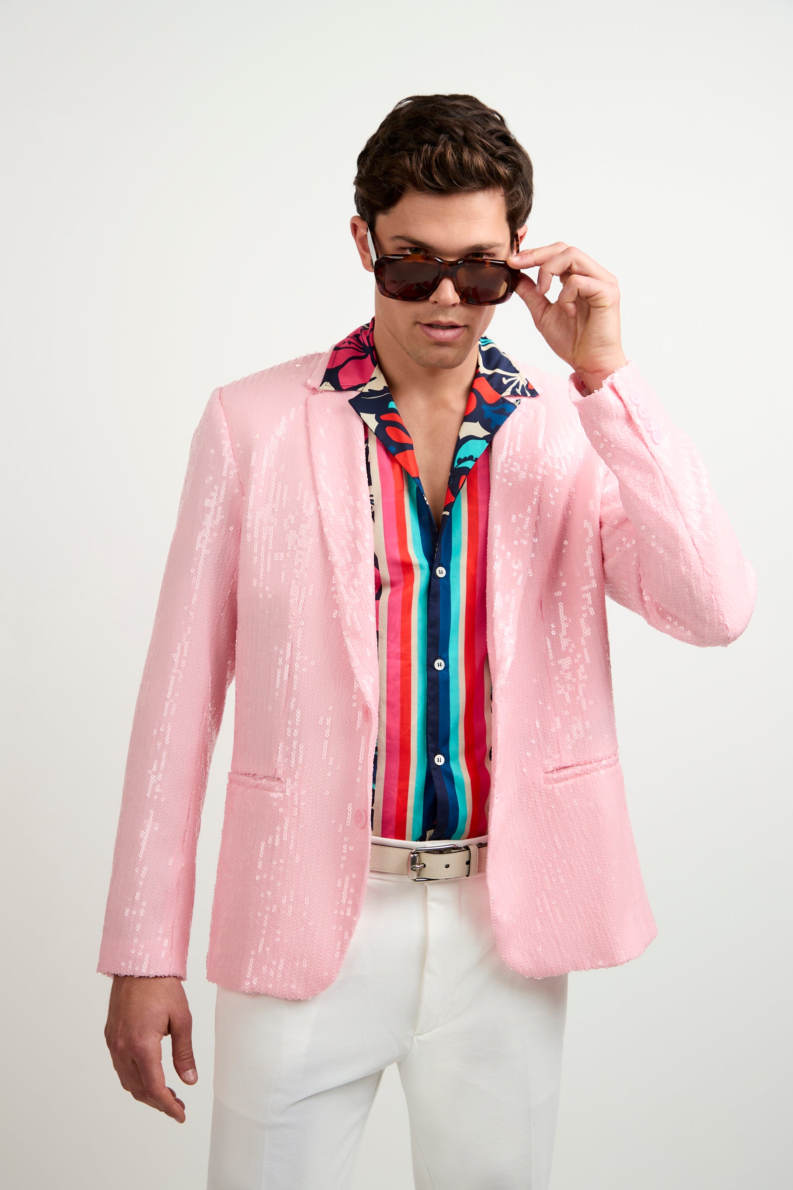 A man in a Bowie Sequin Blazer, colorful striped shirt, white pants, and sunglasses stands against a plain white background, lifting his sunglasses slightly with one hand.
