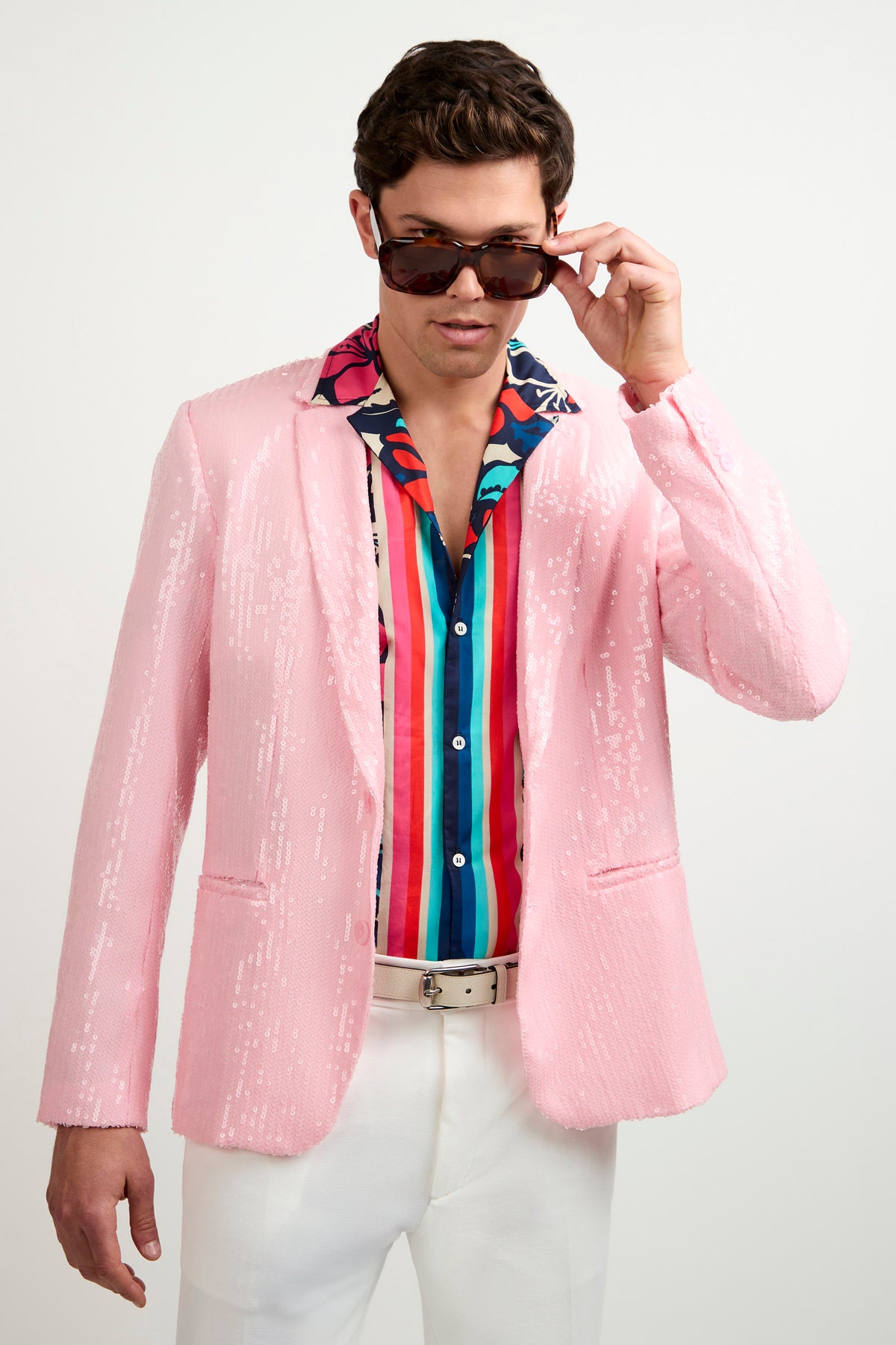 A man wearing the Bowie Sequin Blazer, a vibrant striped shirt, white pants, and sunglasses adjusts his shades while looking down against a plain white background.