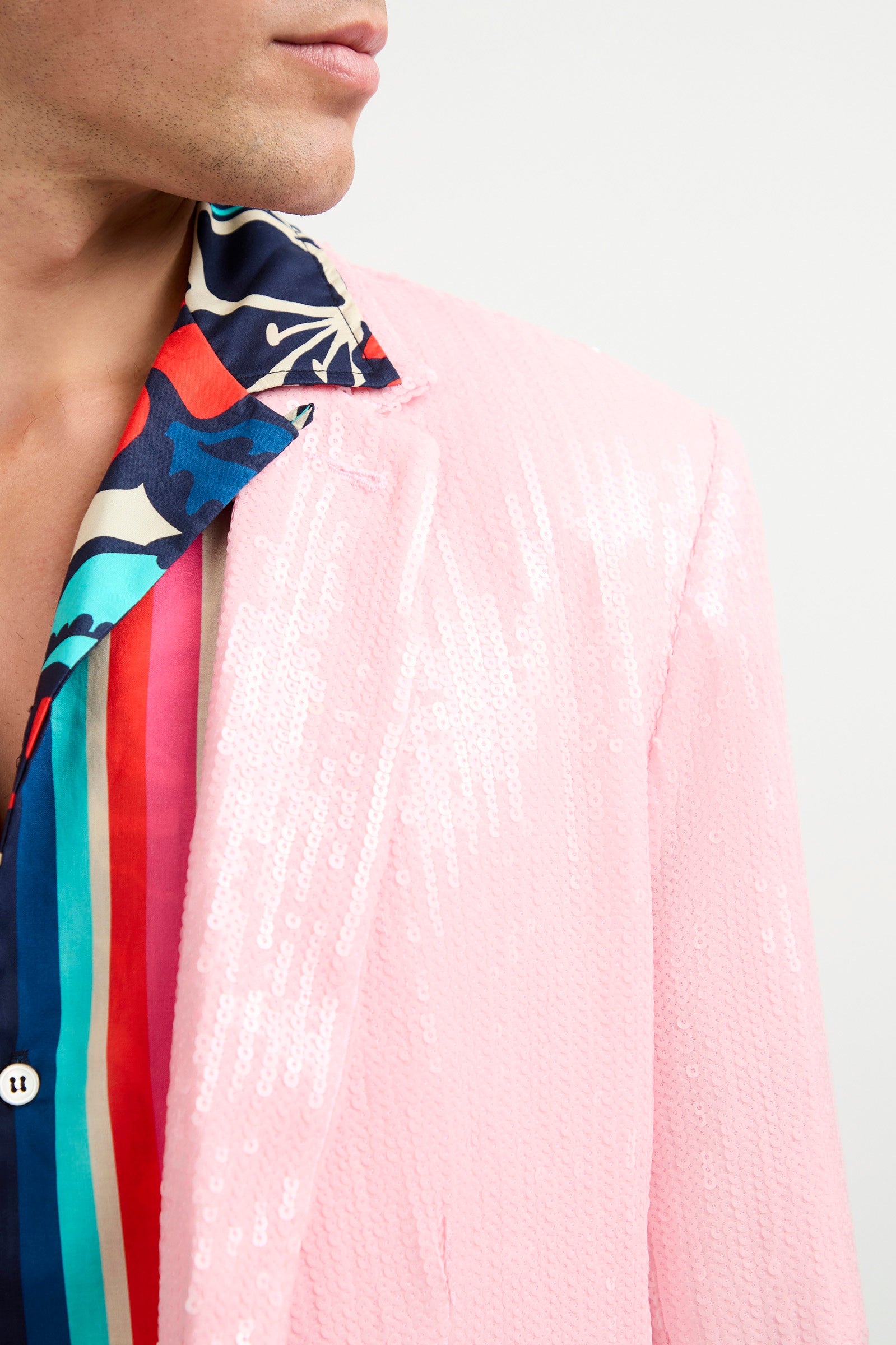A person wears the Bowie Sequin Blazer in shiny light pink over a vibrant, boldly striped and abstract patterned shirt. The image is cropped, showing their face partially turned away.